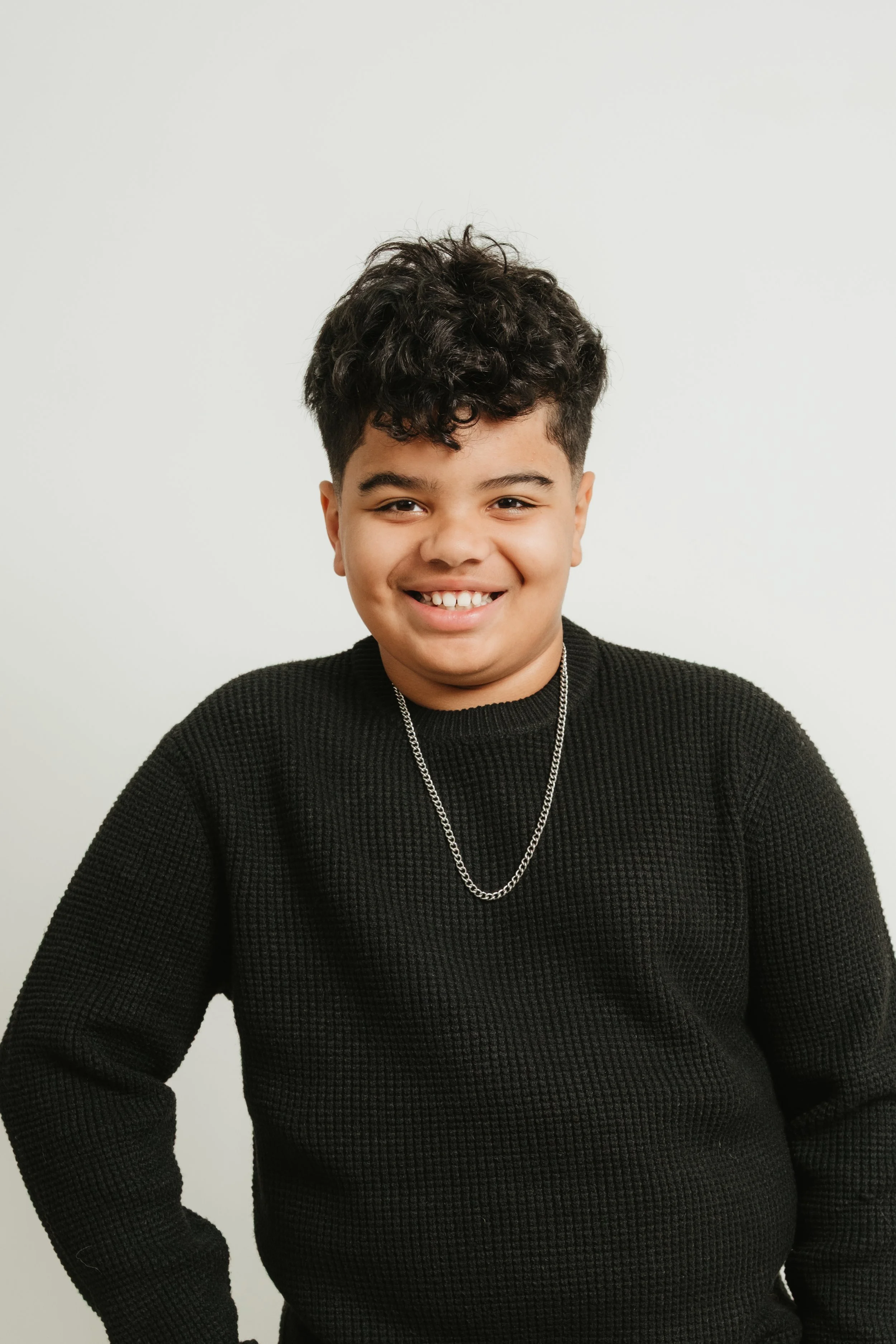 Portrait of a smiling young boy with curly dark hair, wearing a black sweater and a silver chain necklace, standing against a plain white background.