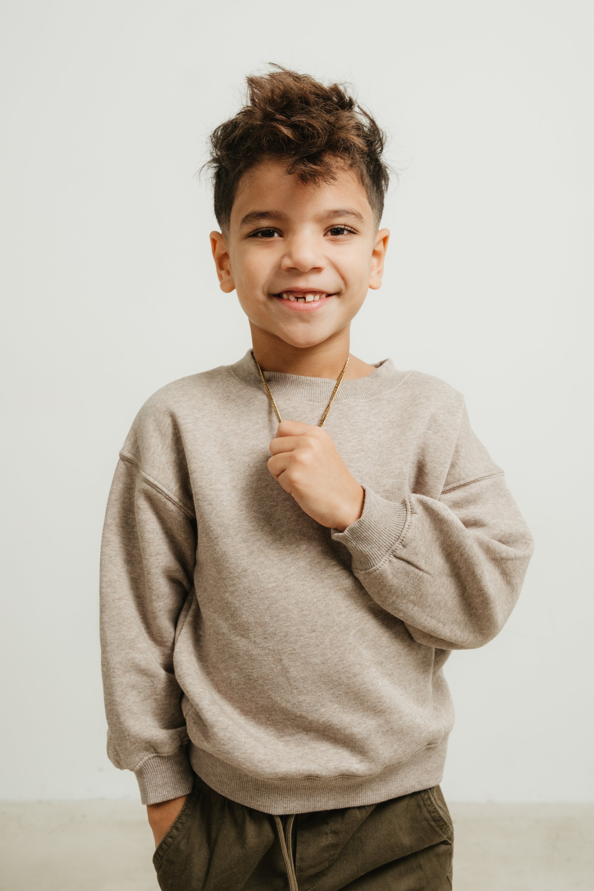A young boy with curly hair smiling and holding a chain necklace, wearing a beige sweatshirt and dark pants, standing against a plain white wall.