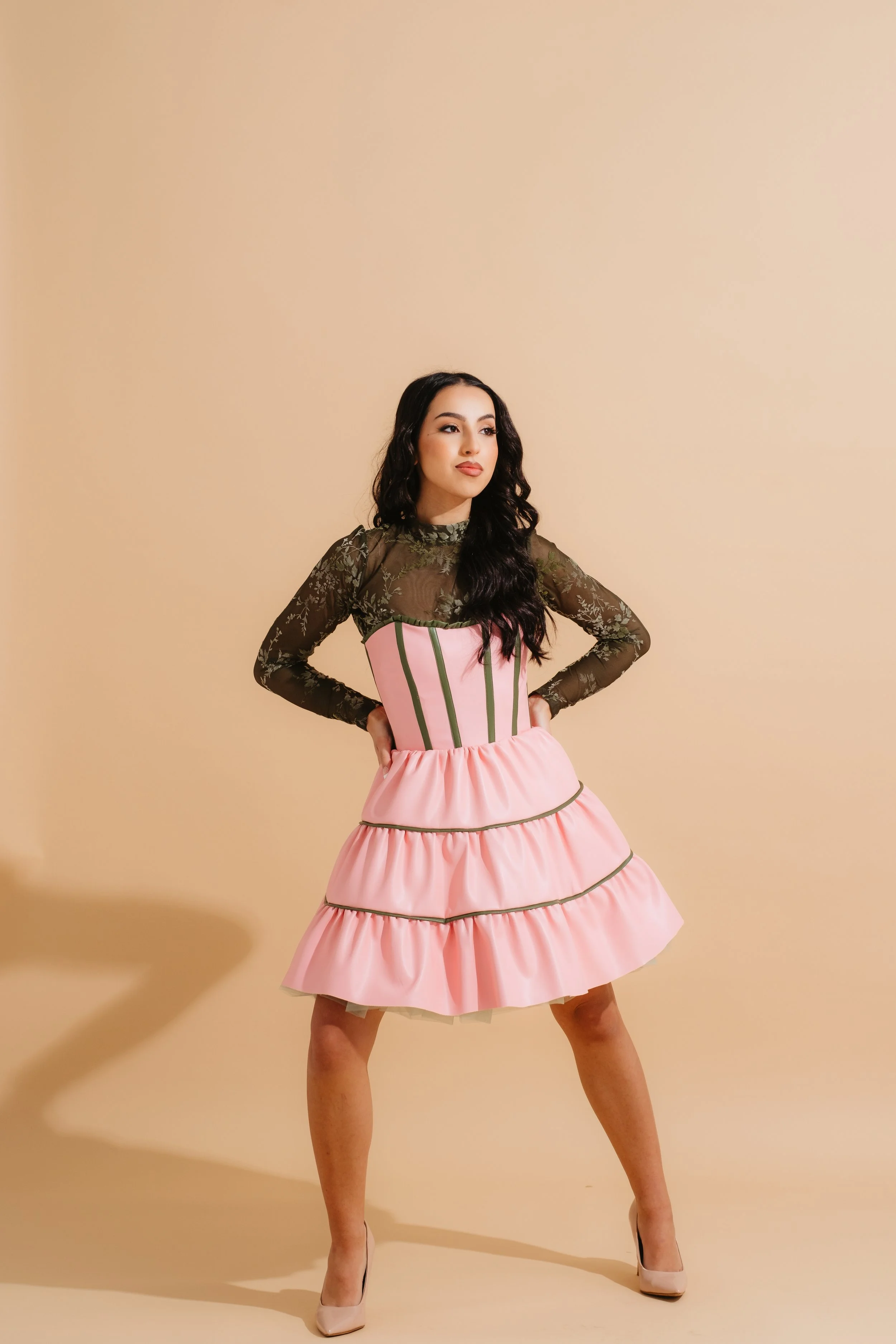 A woman with long dark hair and light skin wearing a dress with a sheer black lace top and a pink tiered skirt, standing with hands on hips against a neutral beige background.
