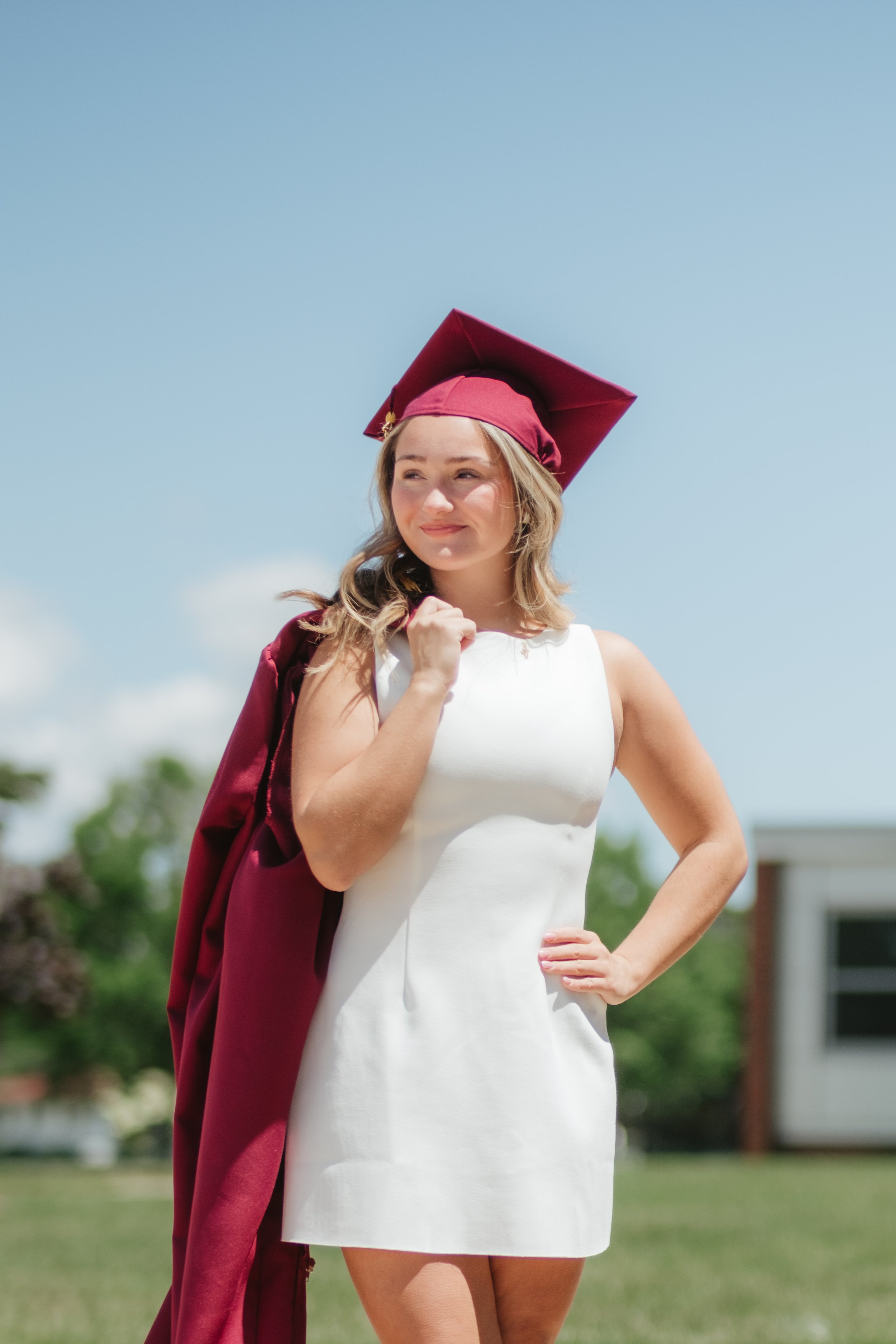 A young woman in a white dress wearing a maroon graduation cap and gown over her shoulder, standing outdoors on a sunny day.