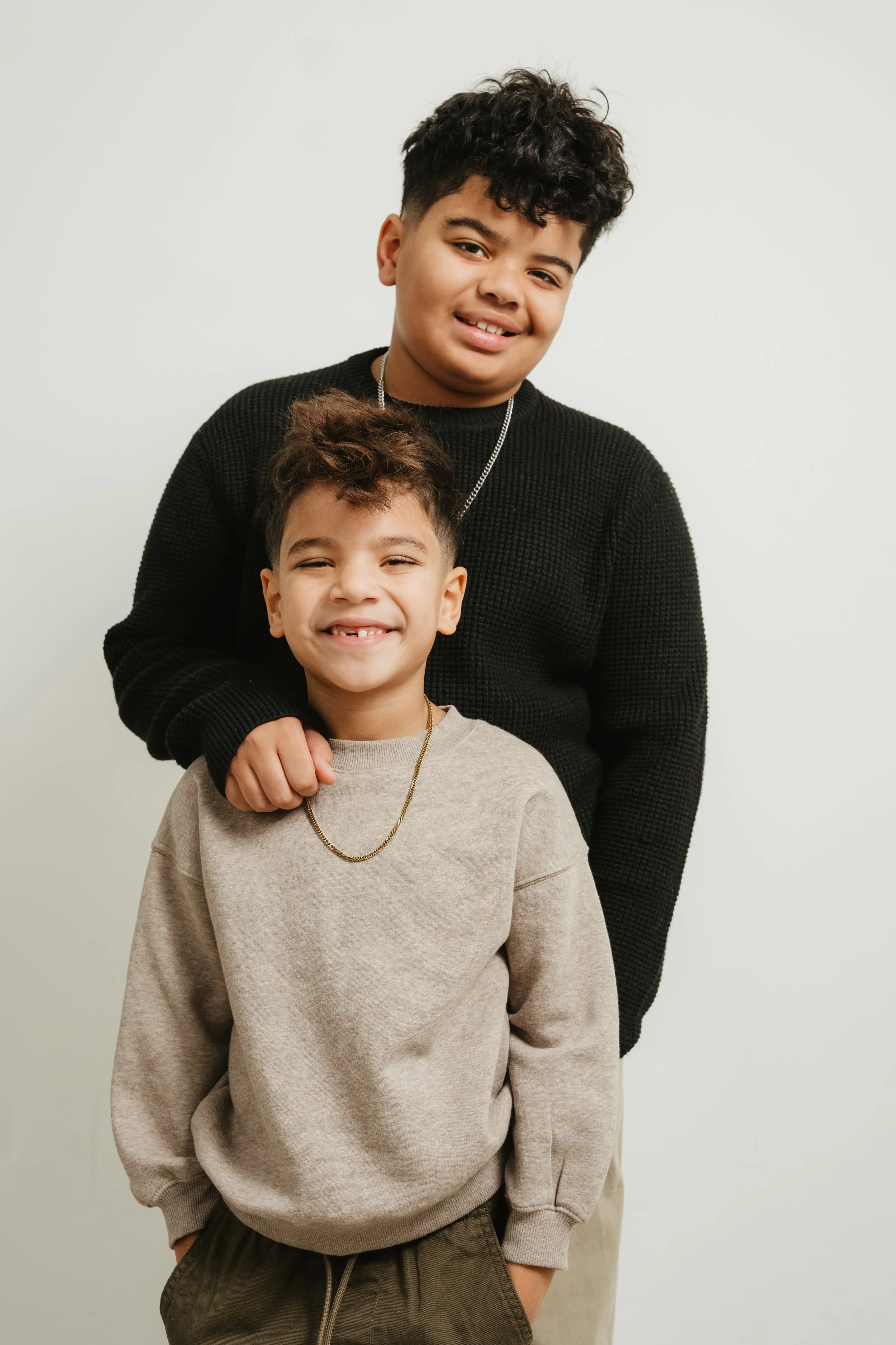 Two boys smiling, with the older boy standing behind the younger, holding the younger's shoulder, against a plain light-colored background.
