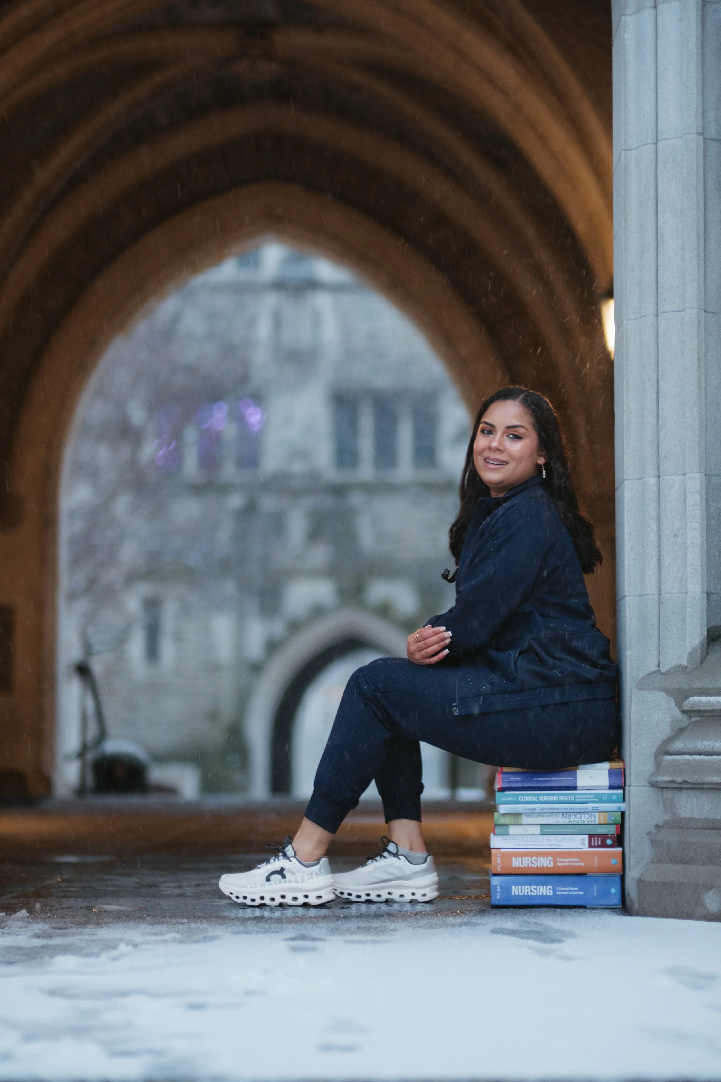 A woman in dark clothing, sitting on a stack of nursing textbooks under an architectural archway, with a rainy city background.