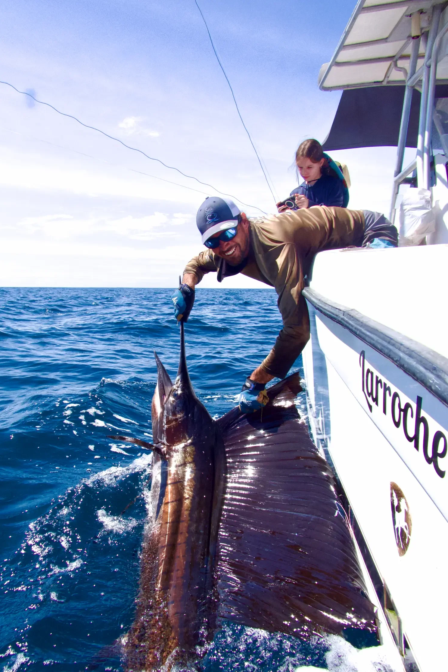Boat captain holding a pacific sailfish while in action during a safe release wearing our sought after merch.