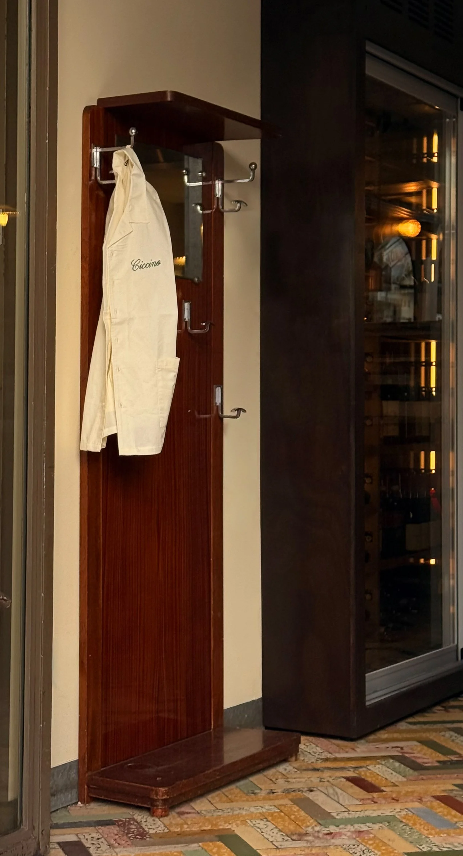 Wooden coat rack with hooks, a white chef's jacket hanging on it, in a restaurant or hotel hallway.