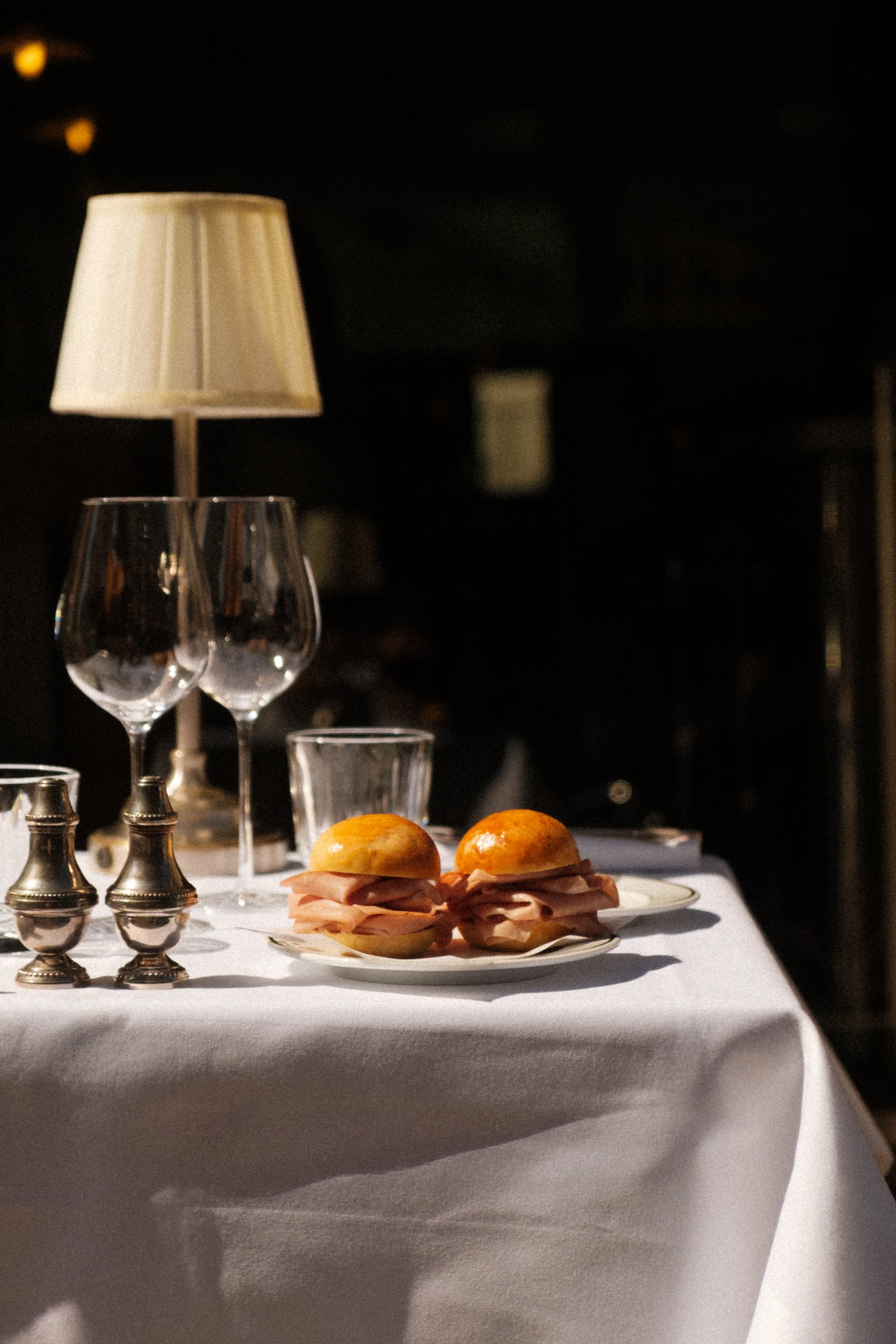 A table set with two ham sandwiches on a white plate, two empty wine glasses, a glass, a pair of silver salt and pepper shakers, and a table lamp with a beige lampshade in a dimly lit room. Architecture d'intérieur, design, concept, deco