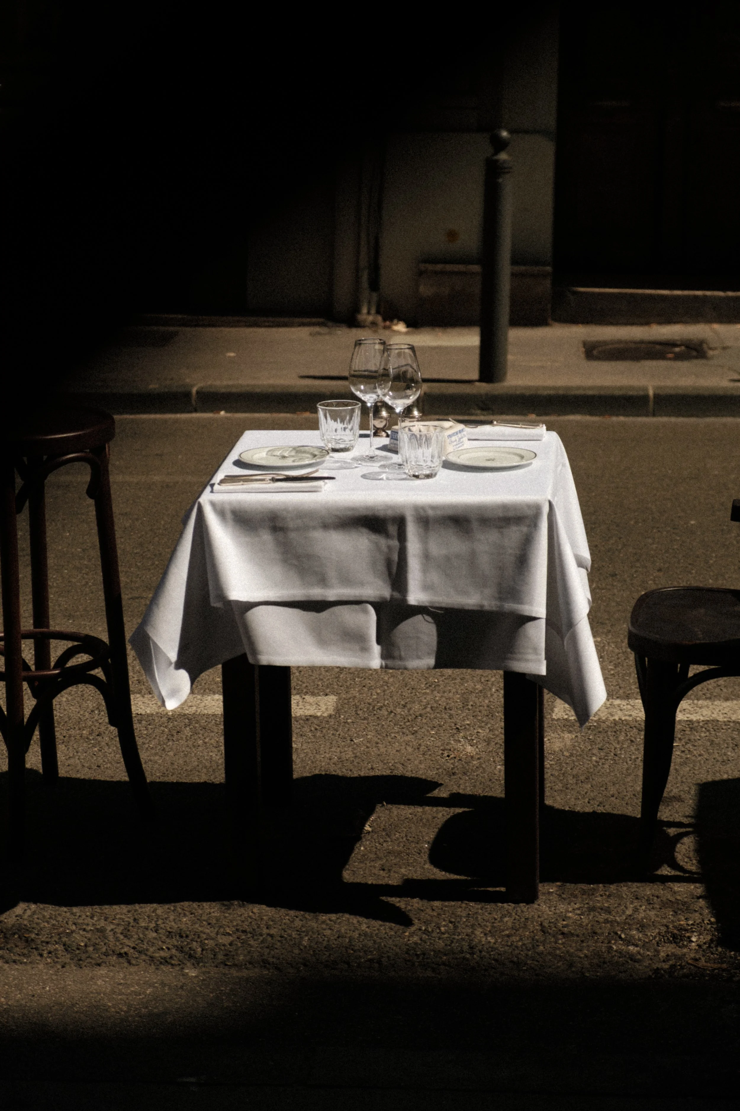 A set dining table with a white tablecloth, wine glasses, water glasses, plates, and silverware, outdoors on a street at night, illuminated by a streetlight.