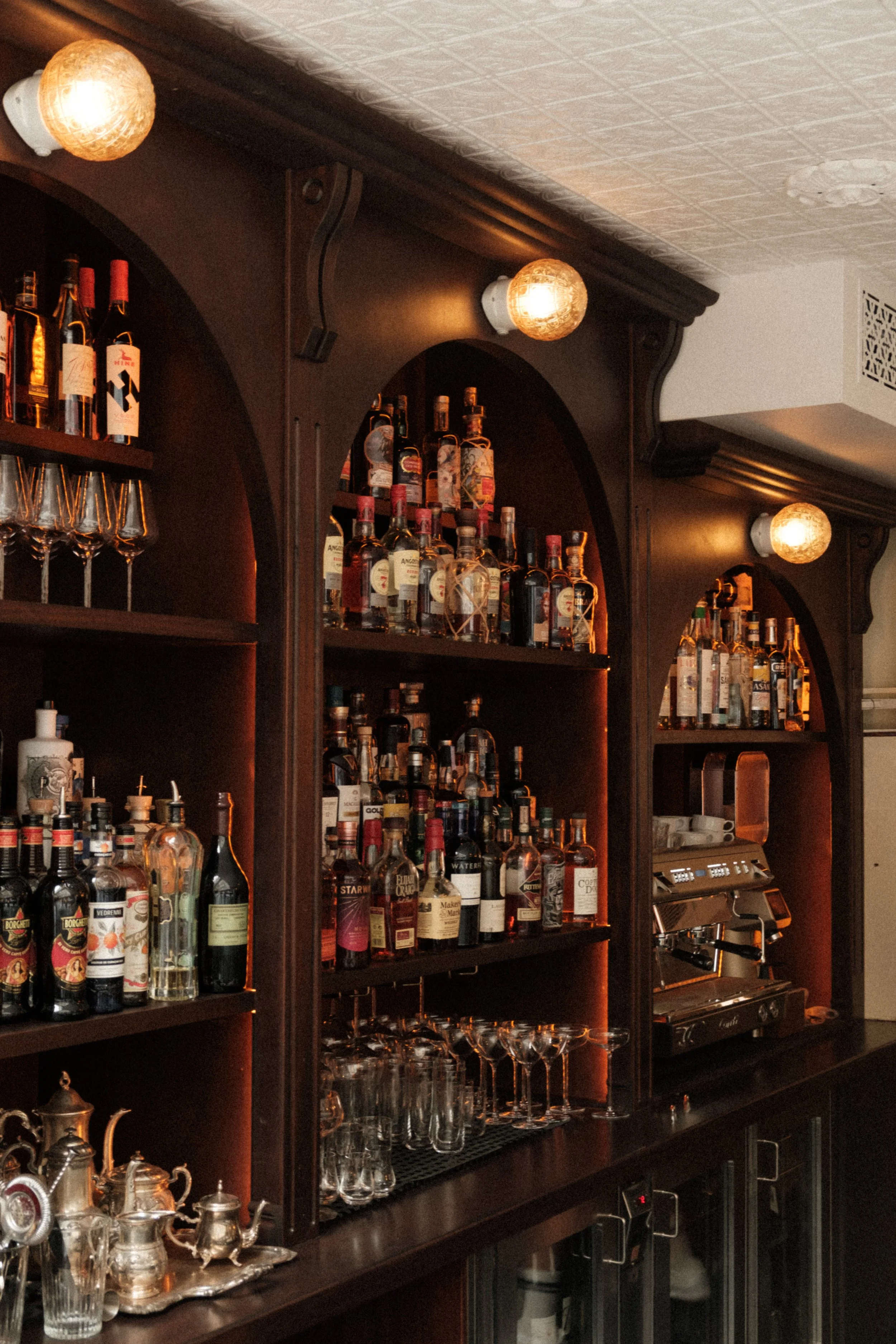 Bar with dark wooden shelves stocked with various bottles of liquor, wine glasses on the counter, and warm lighting fixtures on the ceiling.
