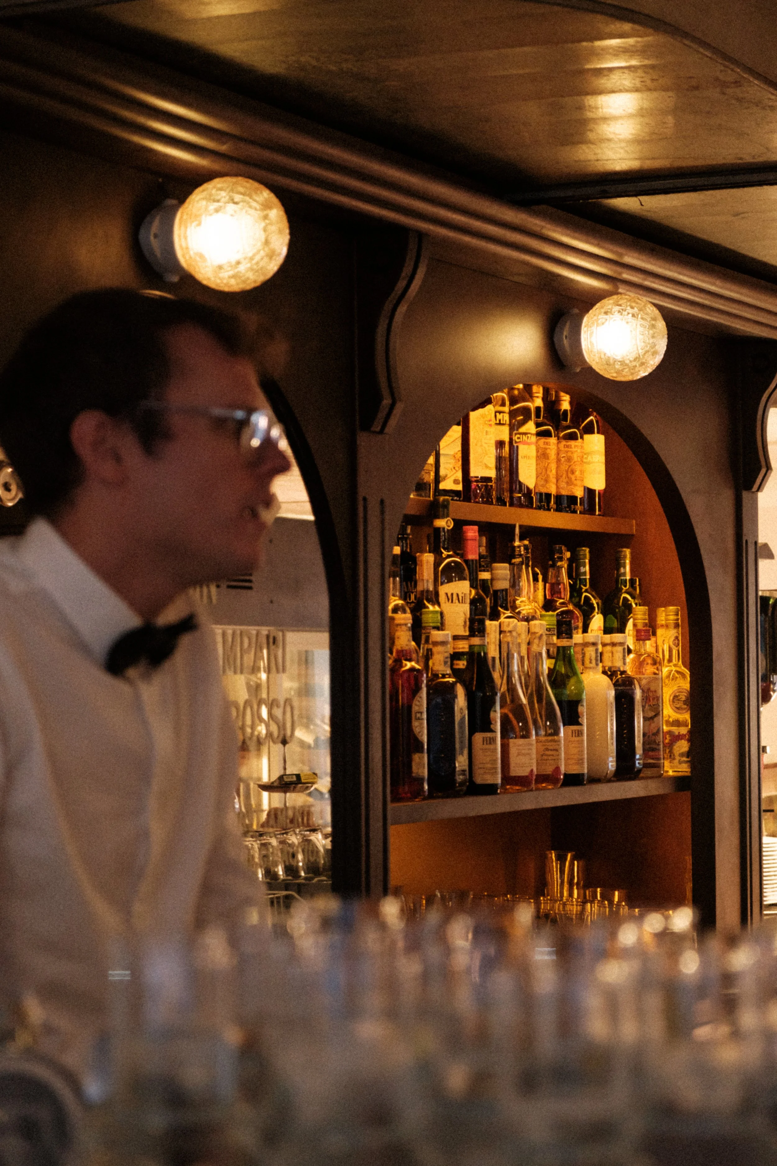 A bartender behind a bar with bottles of alcohol on shelves, dimly lit with two glowing round lights, and a blurred foreground of glasses.