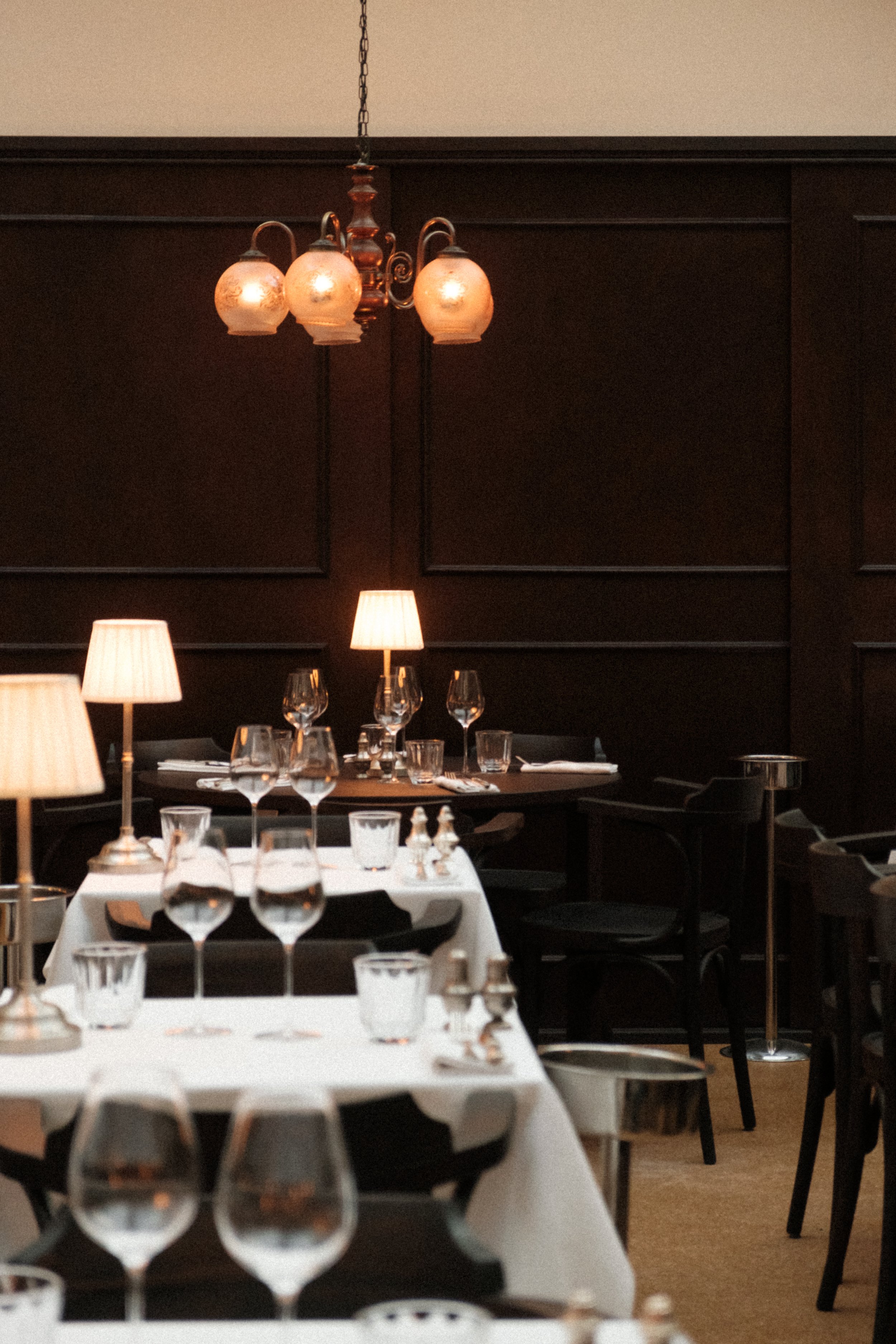 Elegant restaurant interior with white-clothed tables set with wine glasses, water glasses, cutlery, and small lamps, dark wood paneling on walls, hanging light fixtures, and dim lighting creating a warm ambiance.