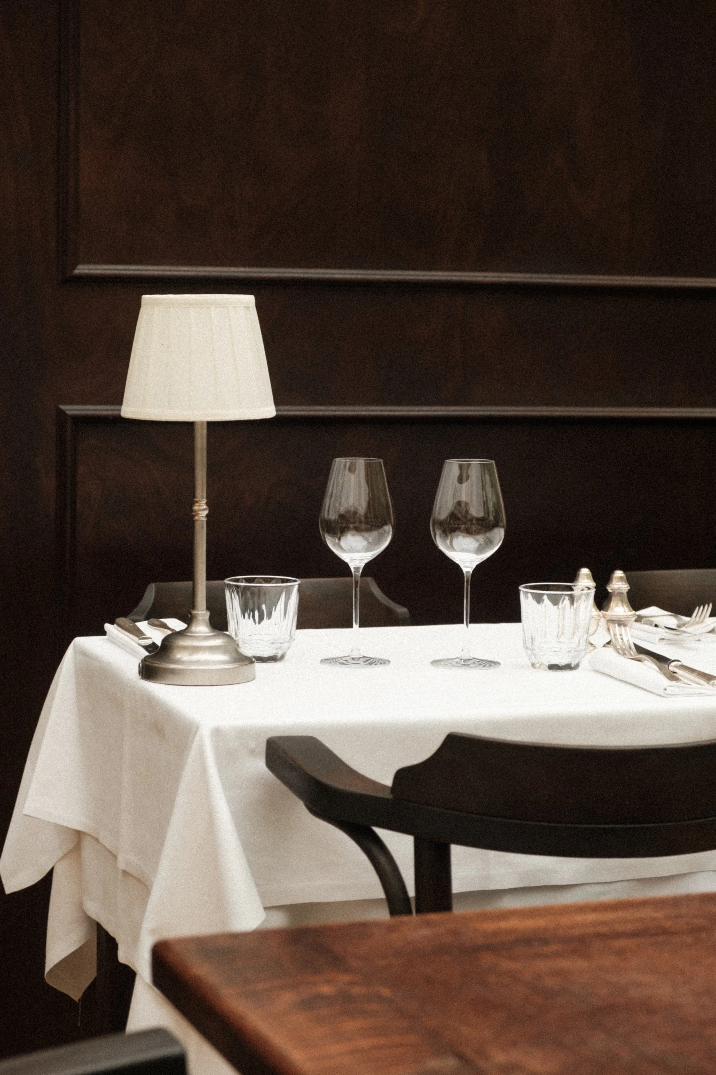Elegant dining table with a white tablecloth, two empty wine glasses, water glasses, a small lamp, and cutlery set in a restaurant setting with dark wood walls.