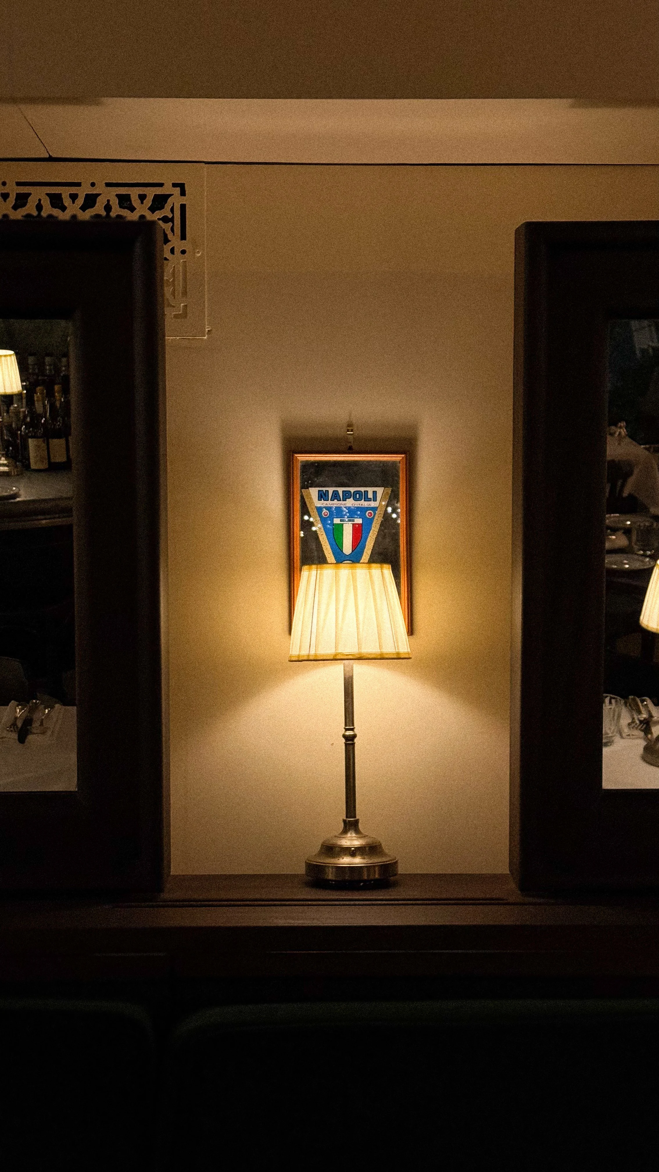 A table lamp with a warm light, positioned in front of a framed football club logo for Napoli, on a wall in a dimly lit room.