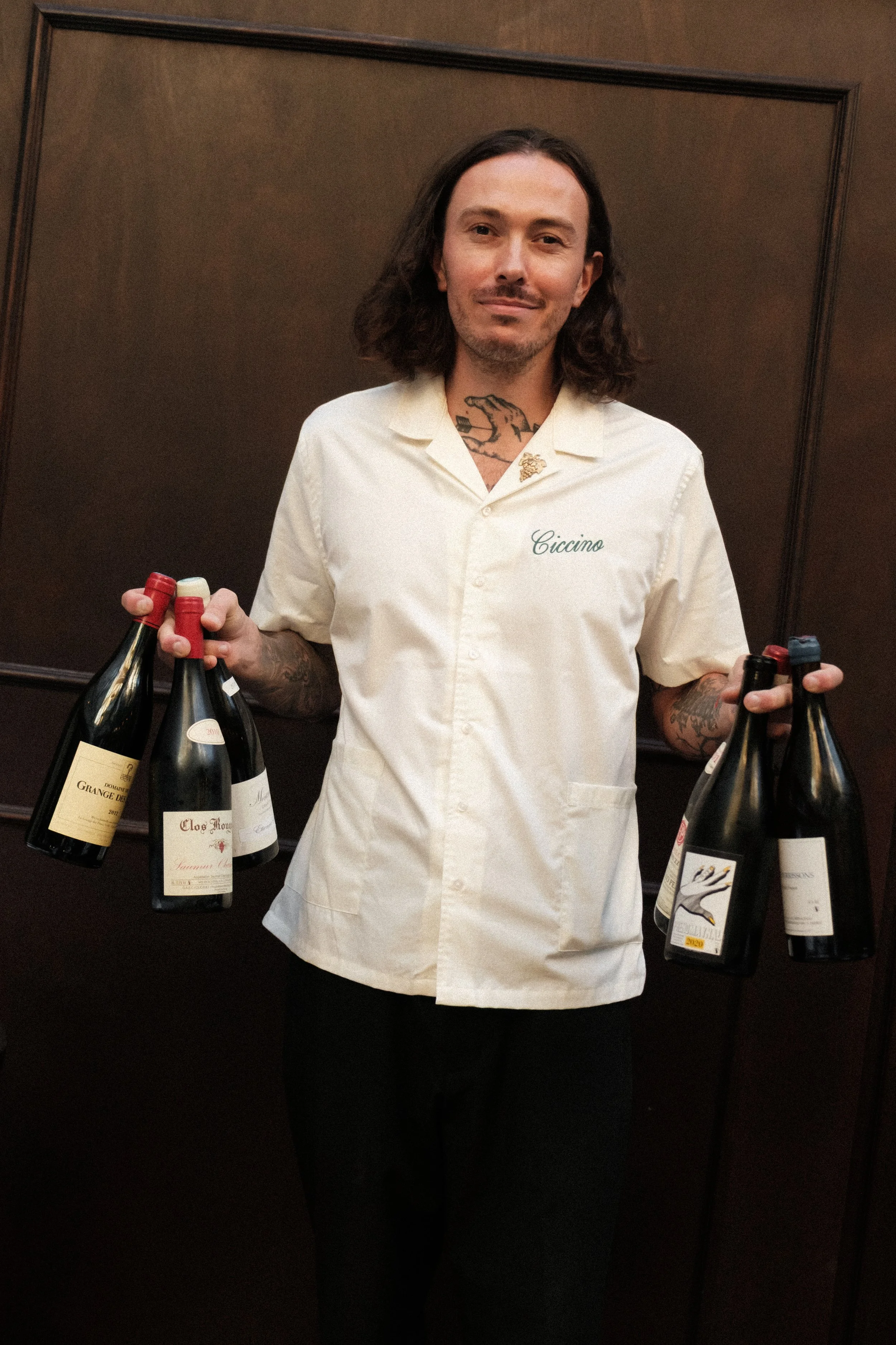 A man with long hair and tattoos on his neck and arms is holding several bottles of wine, standing against a wooden wall.