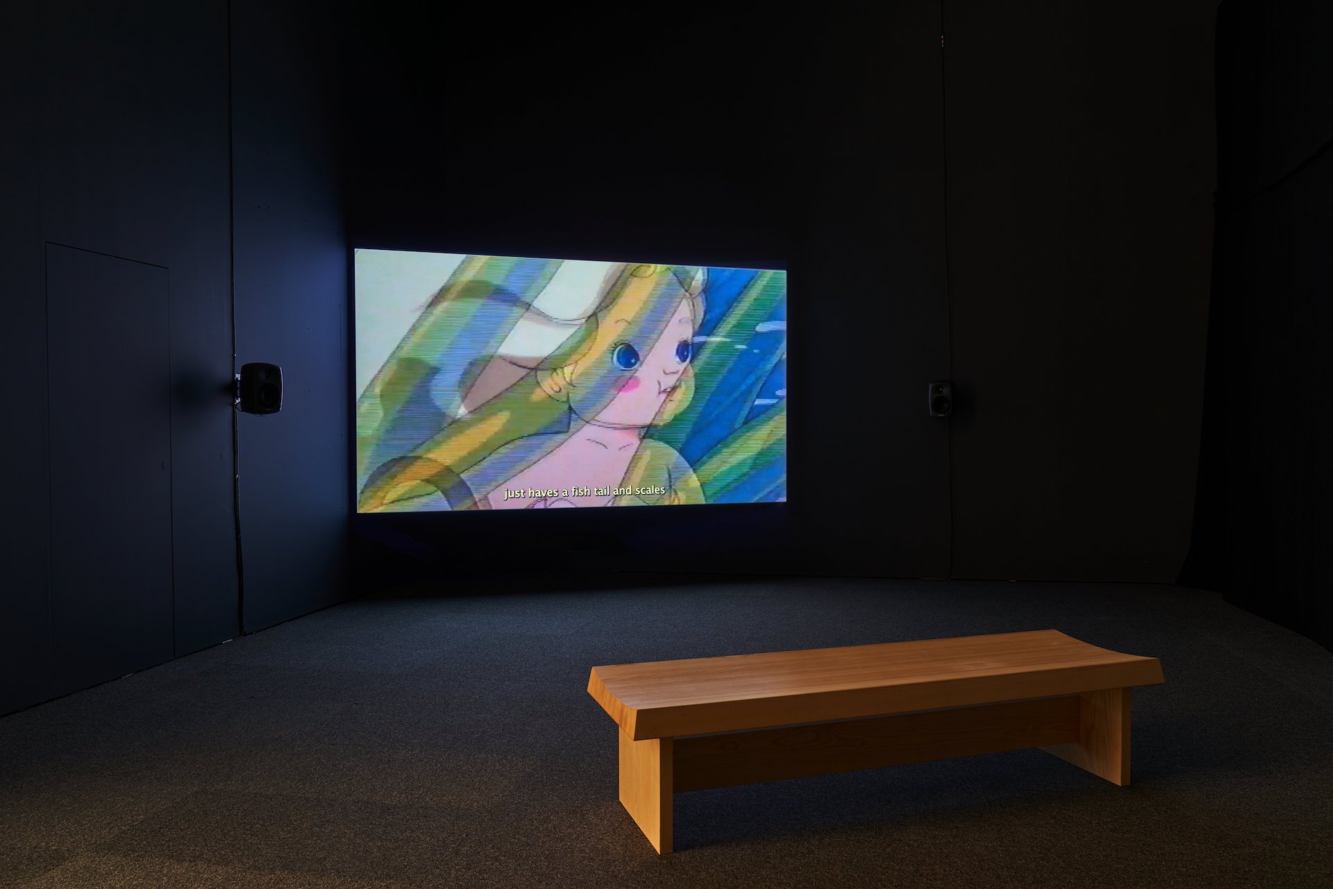  A still image of the asweetsea video installed in a dark room with a wooden bench in front of it. The still image depicts a white cartoon child with long very pale hair, large blue eyes and light pink-toned skin. Semi-translucent sea plants curve in