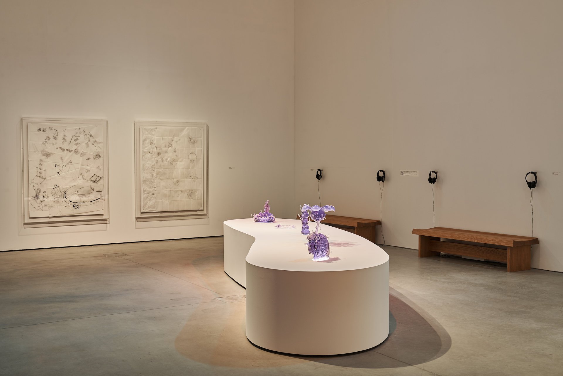  13 - A view of the  asweetsea  exhibition which shows a long, low, curved, white plinth with five purple tinted sculptures evenly spaced on top. The length of the plinth is aligned with the camera angle. Behind the plinth, on the left, are two large