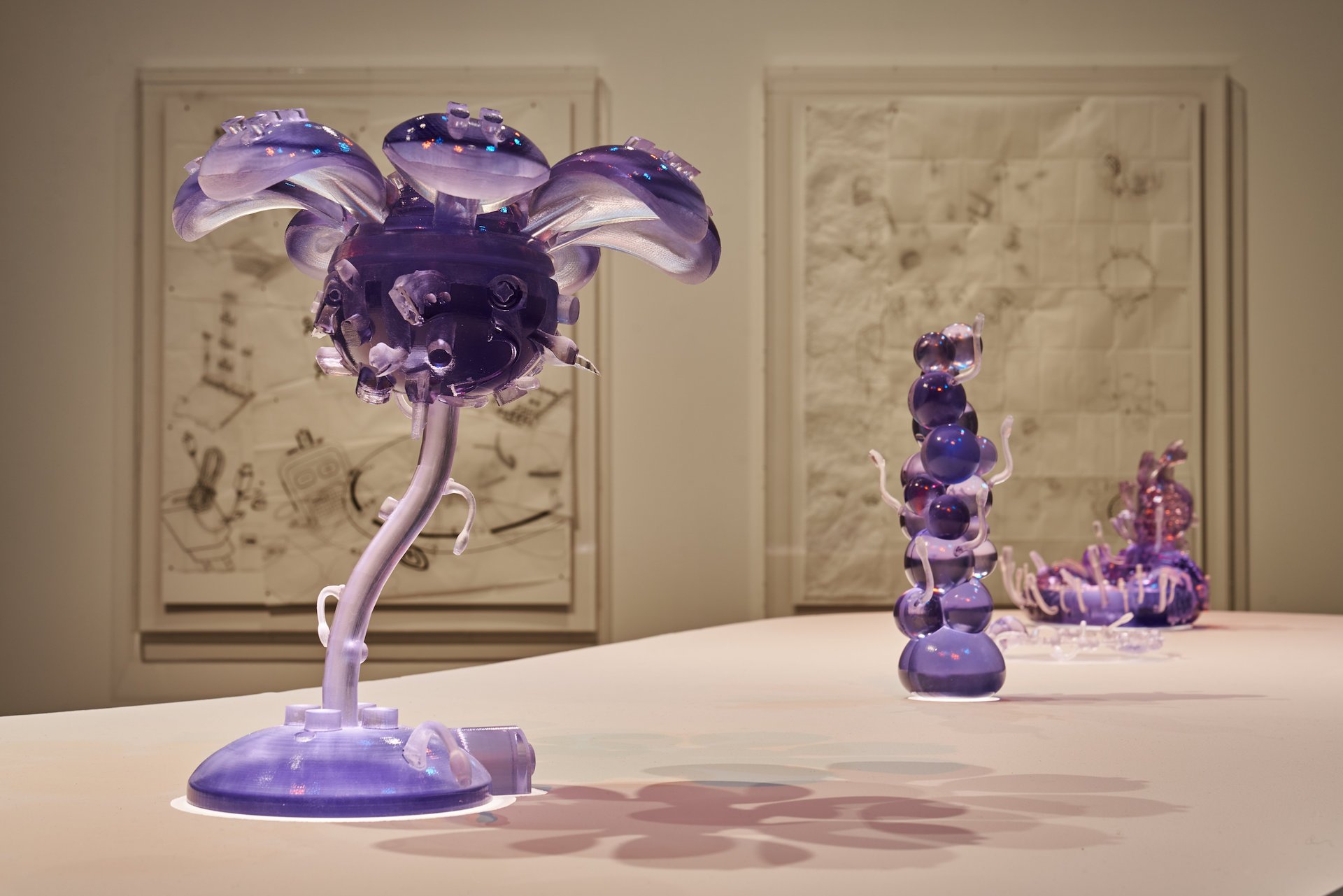  74 - An image which shows the top of a white, curved plinth. On it are four objects which have been 3D printed out of a purple-tinted, semi-transparent resin. Hidden within the plinth and lined up with the base of each object is a light source. The 