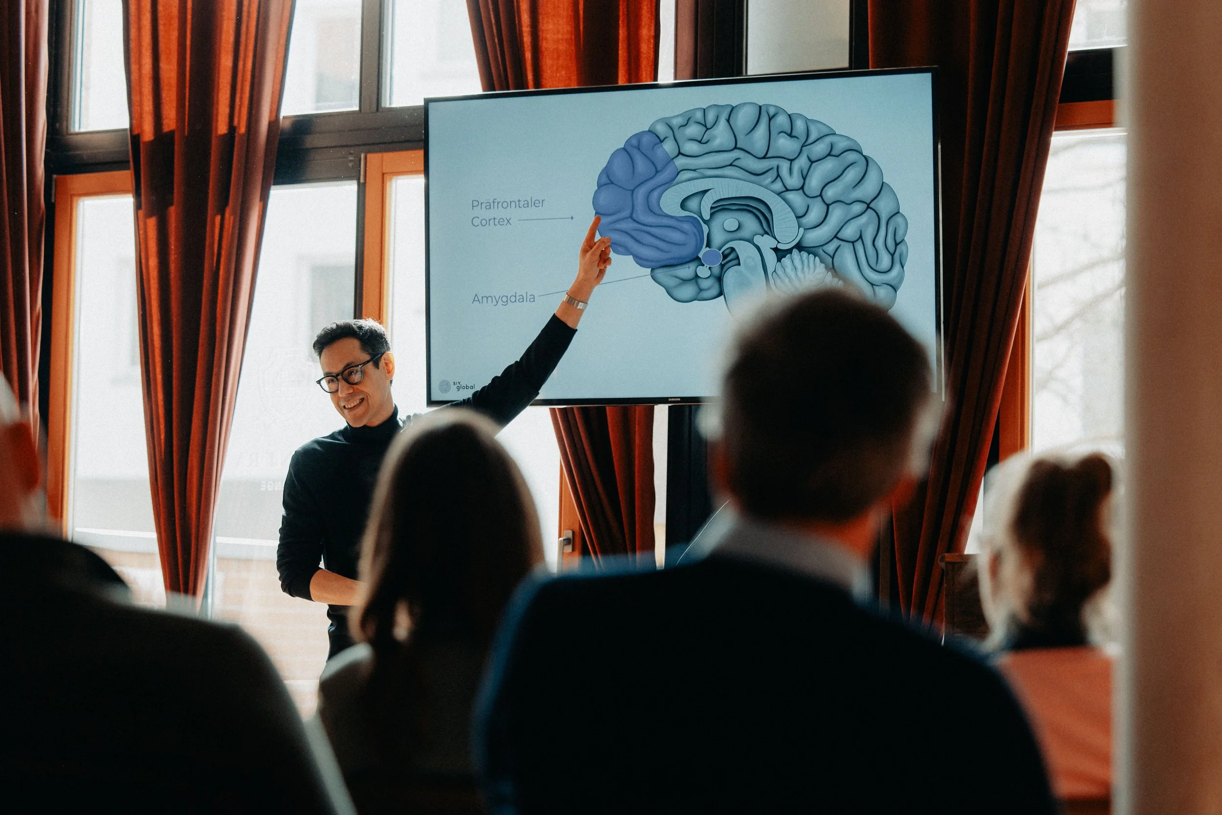 Ein Mann hält eine Präsentation vor einer Gruppe von Menschen, zeigt auf ein Diagramm des Gehirns auf einem Bildschirm, im Raum mit großen Fenstern und roten Vorhängen.