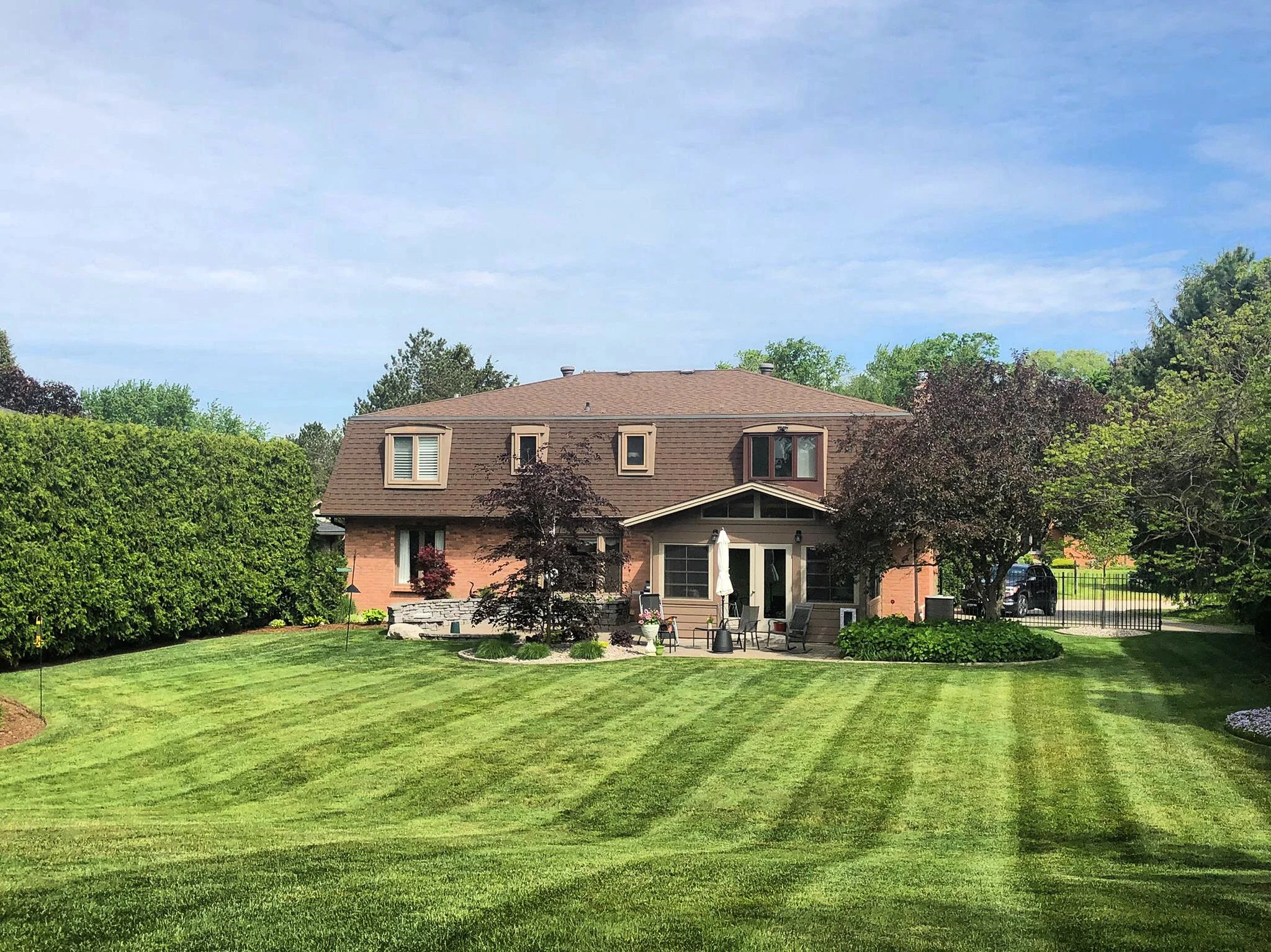 A house with a brown roof and brick exterior, surrounded by a well-maintained lawn and trees, under a partly cloudy sky.