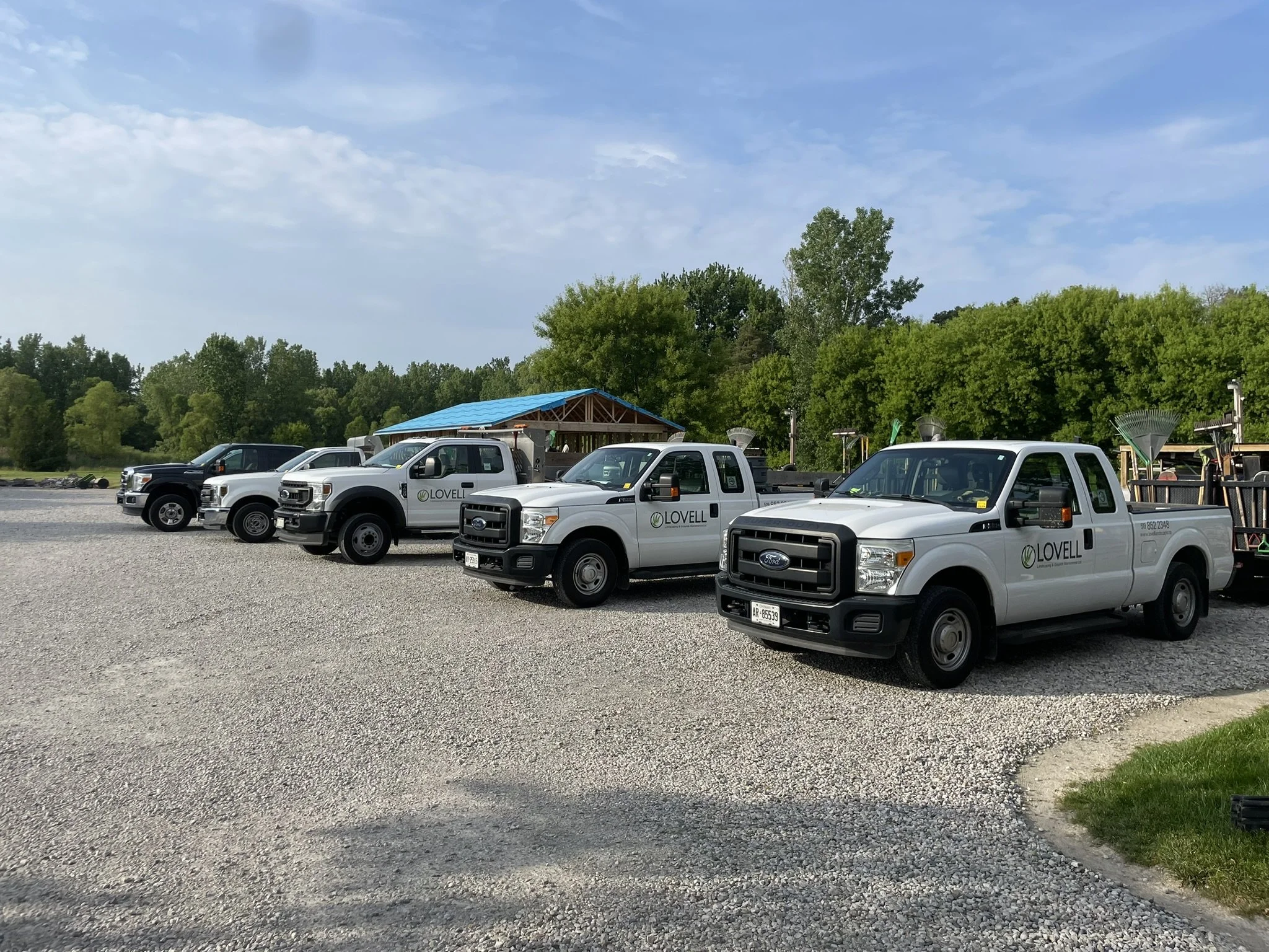 A row of five white pickup trucks with "Lovell" logos parked on a gravel lot, with trees and a blue sky in the background.