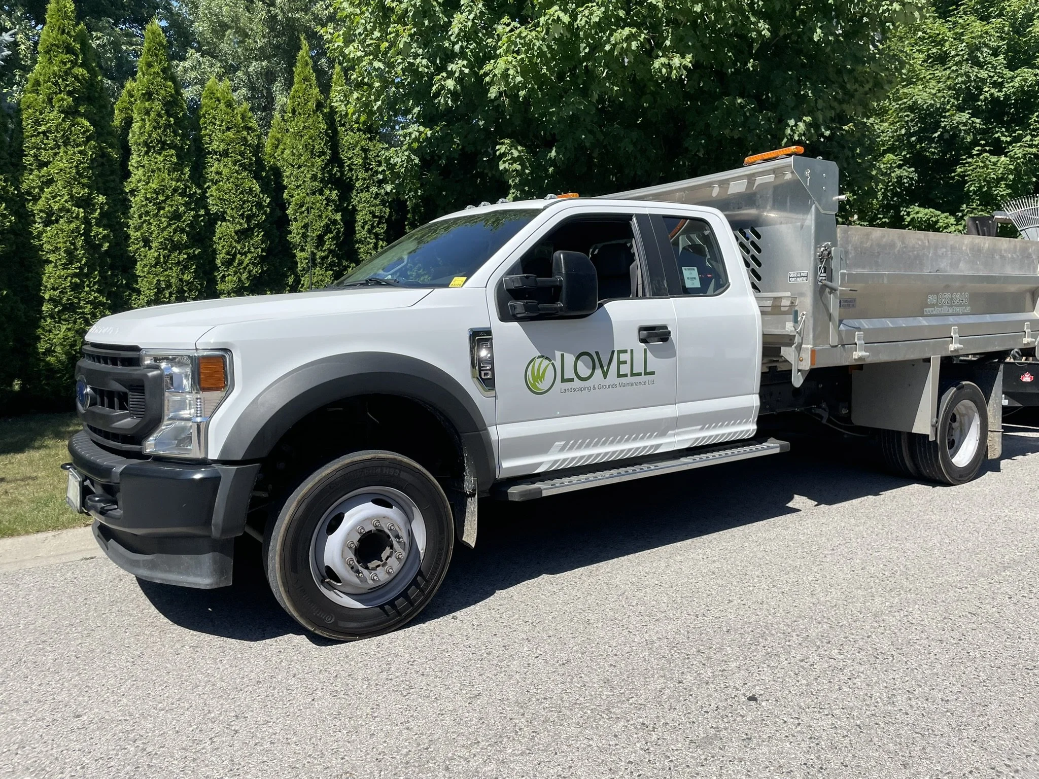 A white Lovell Landscaping and Grounds Maintenance truck parked on a suburban street with green trees in the background.