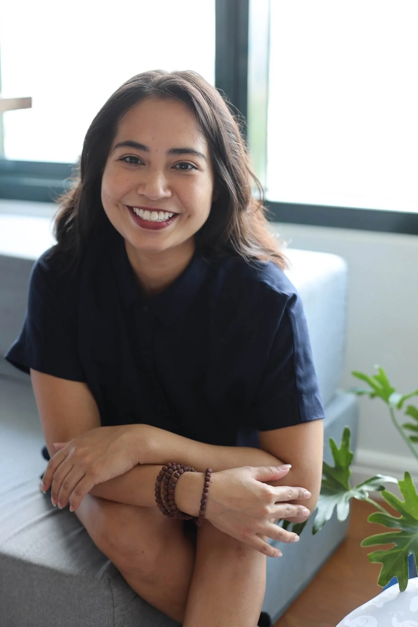 Smiling woman seated on a couch in a bright, modern space, wearing a dark top with arms crossed comfortably, natural light from a window behind her and a green plant nearby.