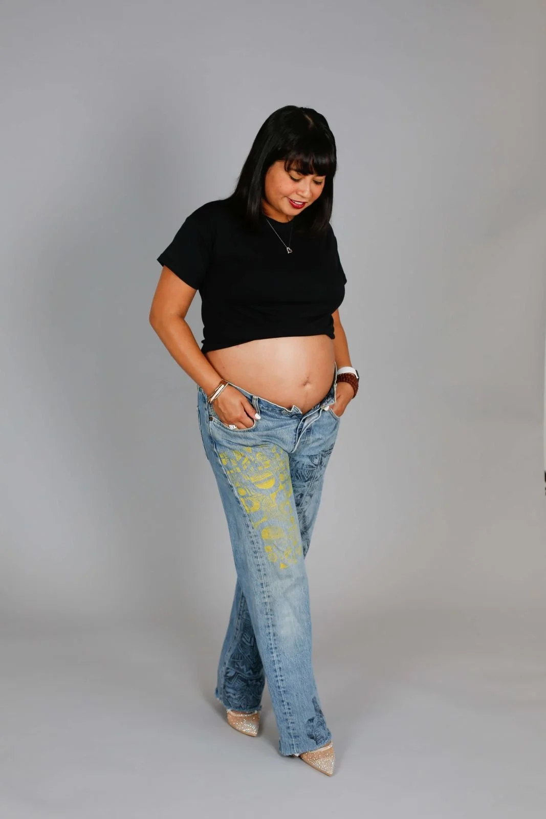 Pregnant woman standing against a neutral background, wearing a black top and jeans, gently looking down at her baby bump in a calm, reflective pose.