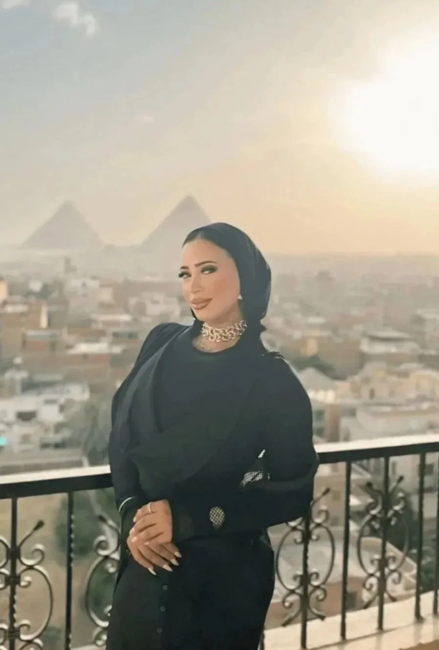 A woman standing on a balcony with a cityscape in the background, including the pyramids of Egypt visible in the distance, during sunset.