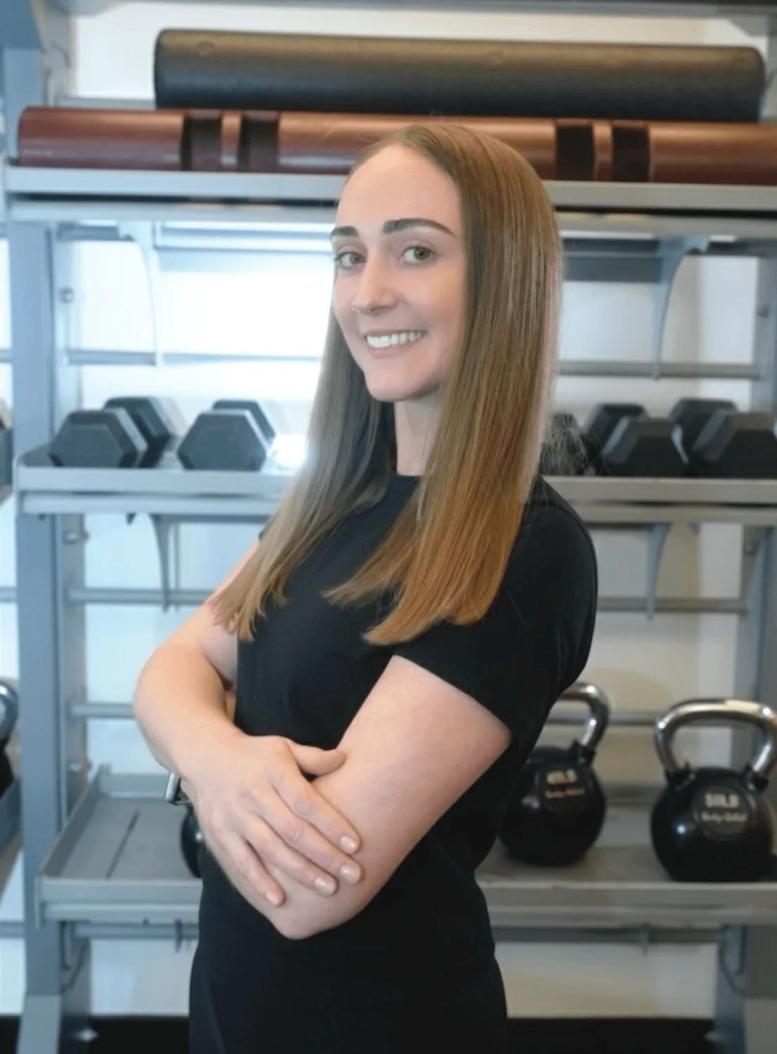 Woman standing in a gym in front of a rack of dumbbells and kettlebells, smiling with arms crossed, wearing a black top.