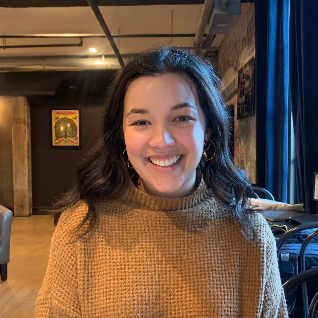 A woman with dark wavy hair, wearing a brown knit sweater, smiling at the camera in a cozy indoor setting with brick walls and artwork in the background.