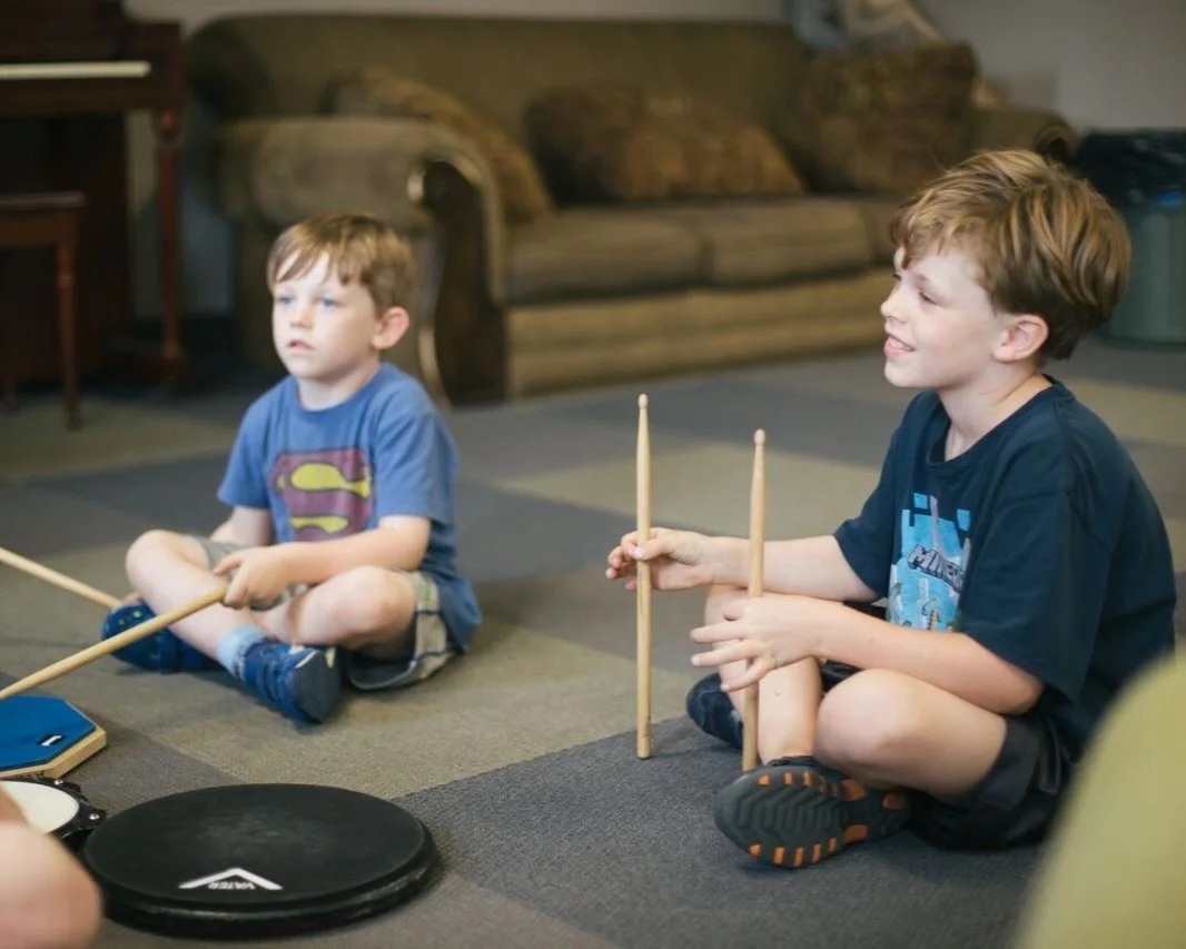 Two young boys sitting on a carpeted floor, holding drumsticks and playing drums. The boy on the right is smiling, and the boy on the left has a serious expression. A couch is in the background.