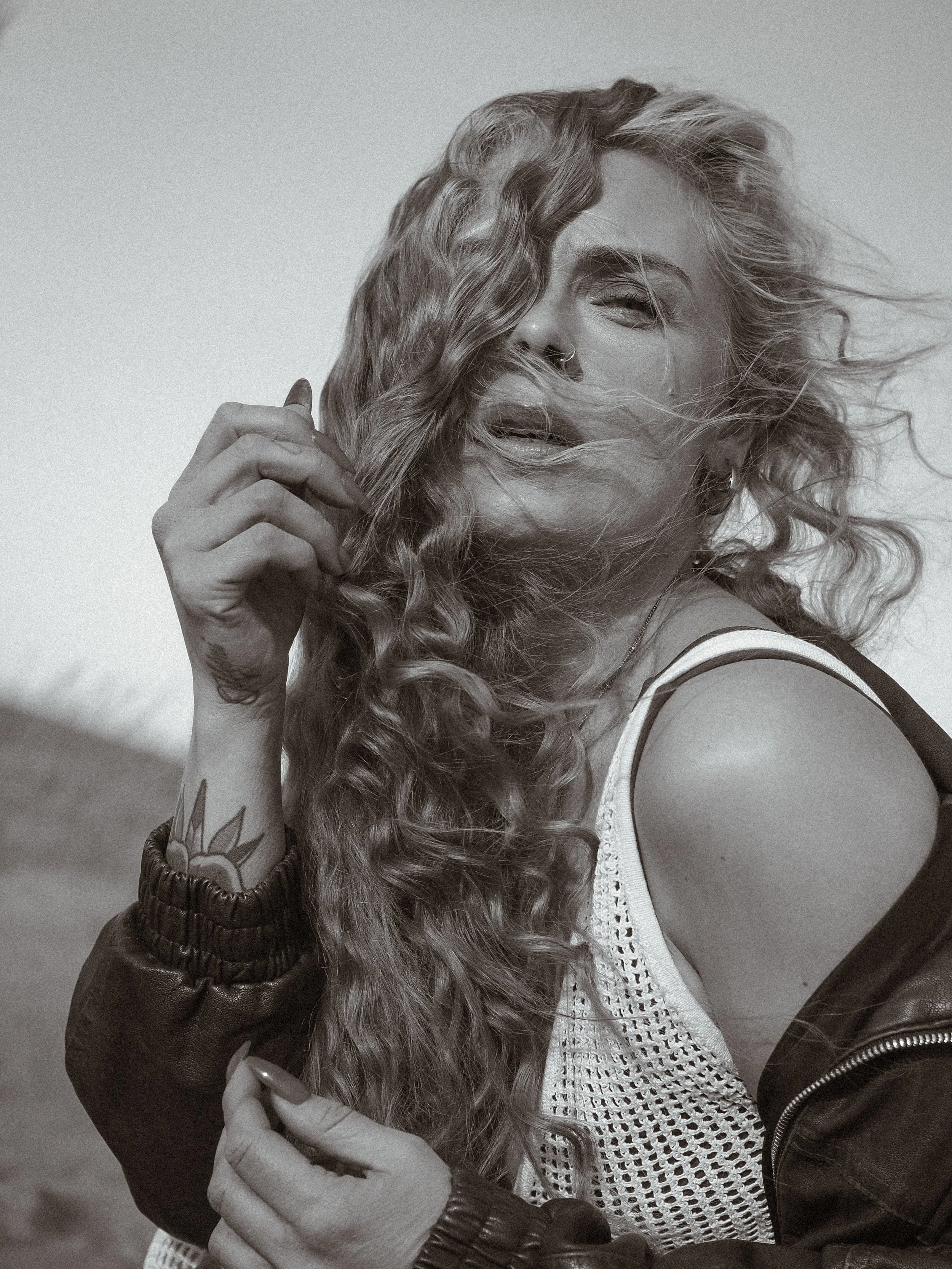 Nuori nainen poseeraa ulkona, hiukset heiluivat tuulessa, hänellä on tatuointeja ja napit nenässä, pukeutuneena valkoiseen neuleeseen ja nahkatakkiin.