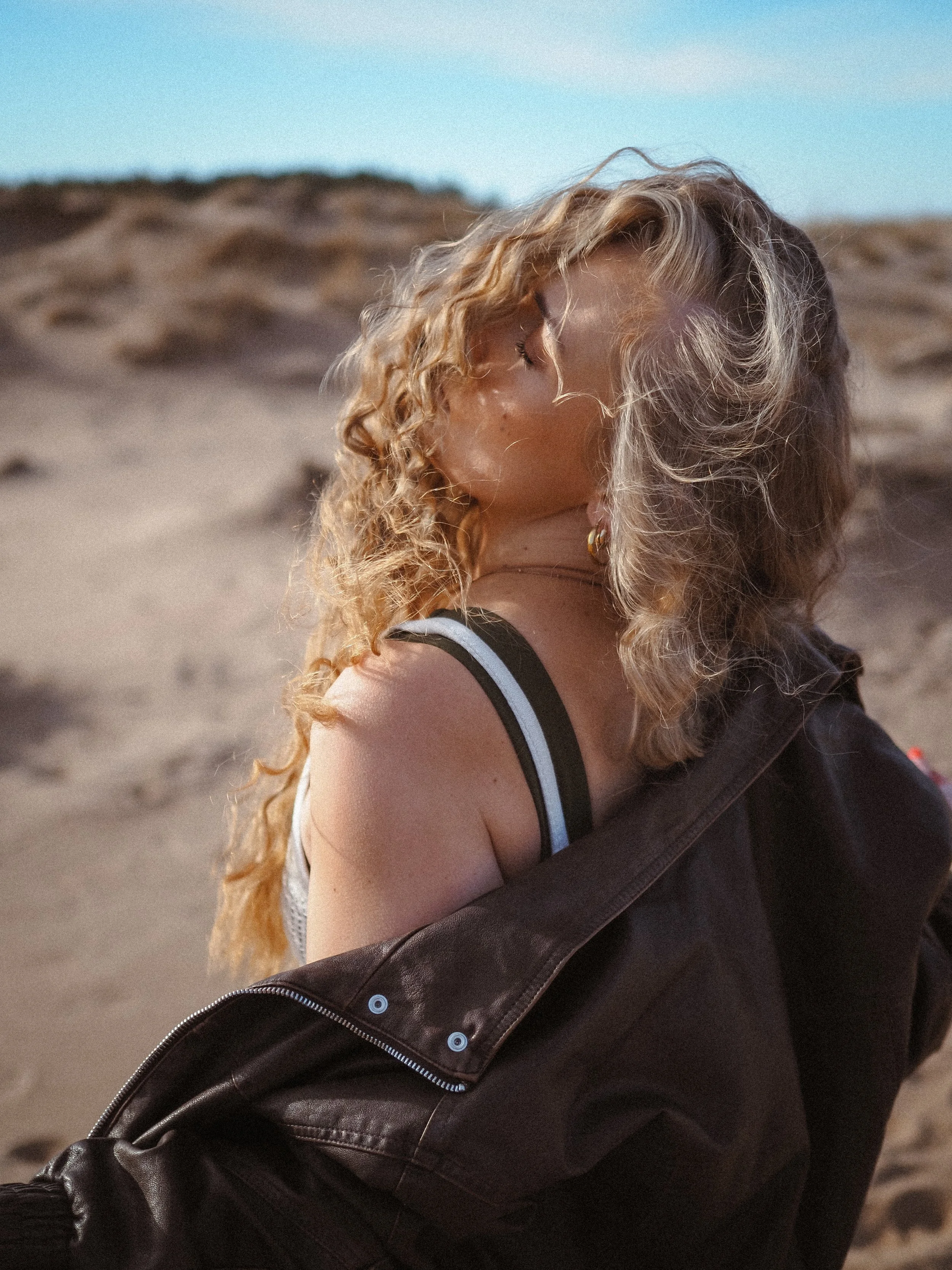 Nuori nainen, jossa kihará hiuksia, istuu rantahiekkällä ja kiinnitetty täyteen asuihin, taustalla hiekkän dyynejä ja taivaskuva.