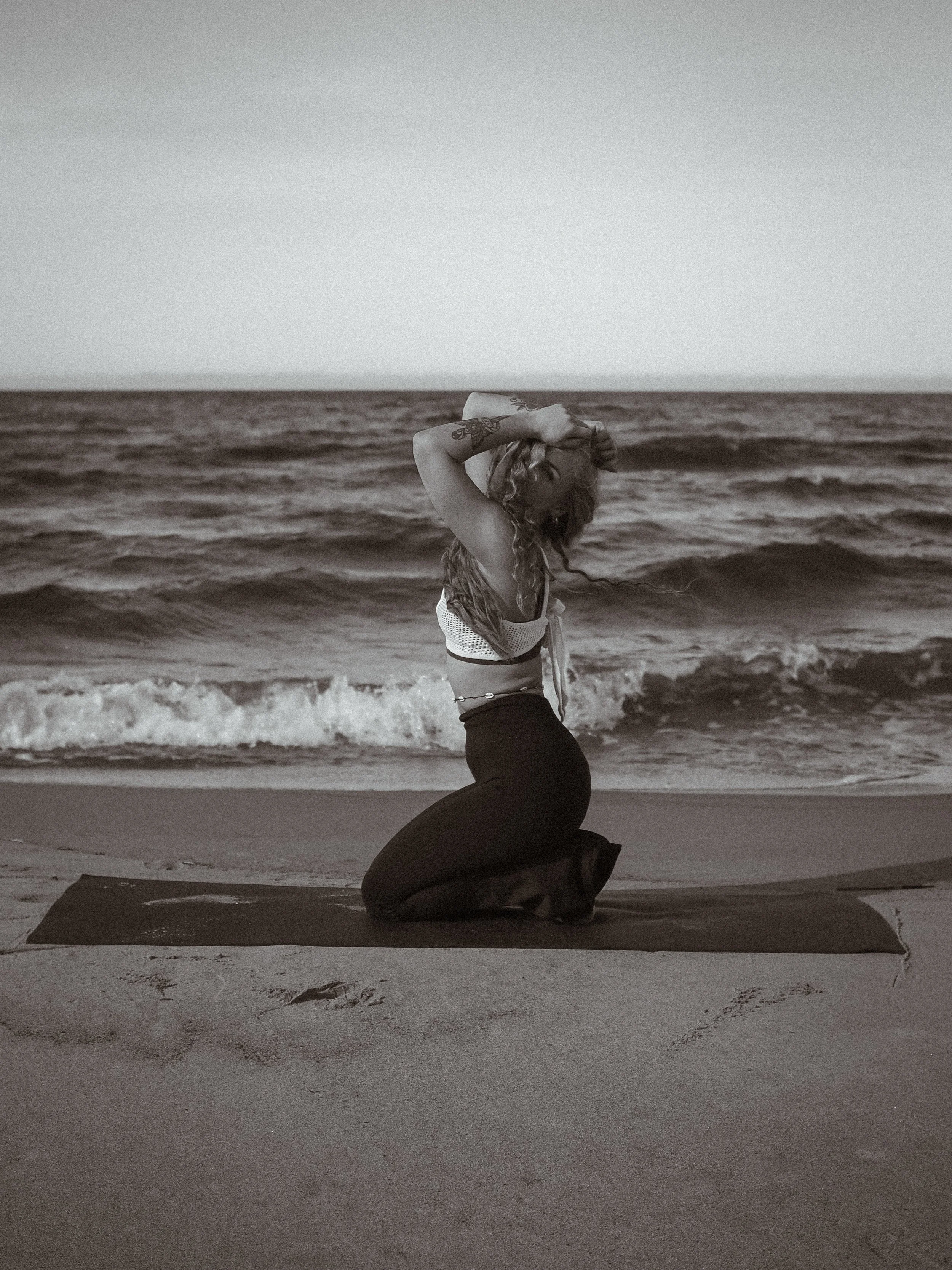 Nainen polvillaan rannalla tekemässä joogaliikettä meren äärellä.