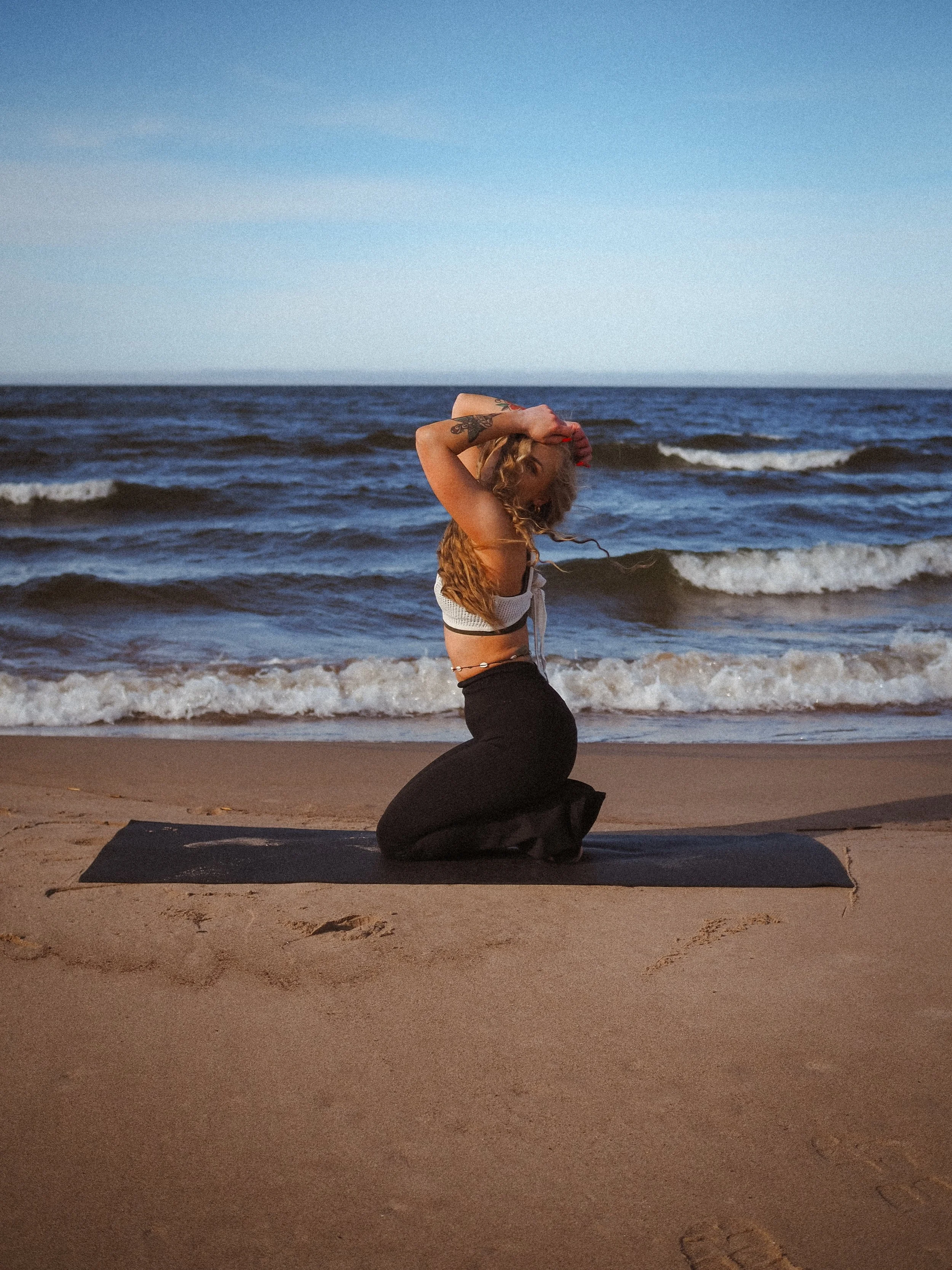 Nainen polvistuu rannalla suorittaen joogaliikettä, taustalla meri ja taivas.