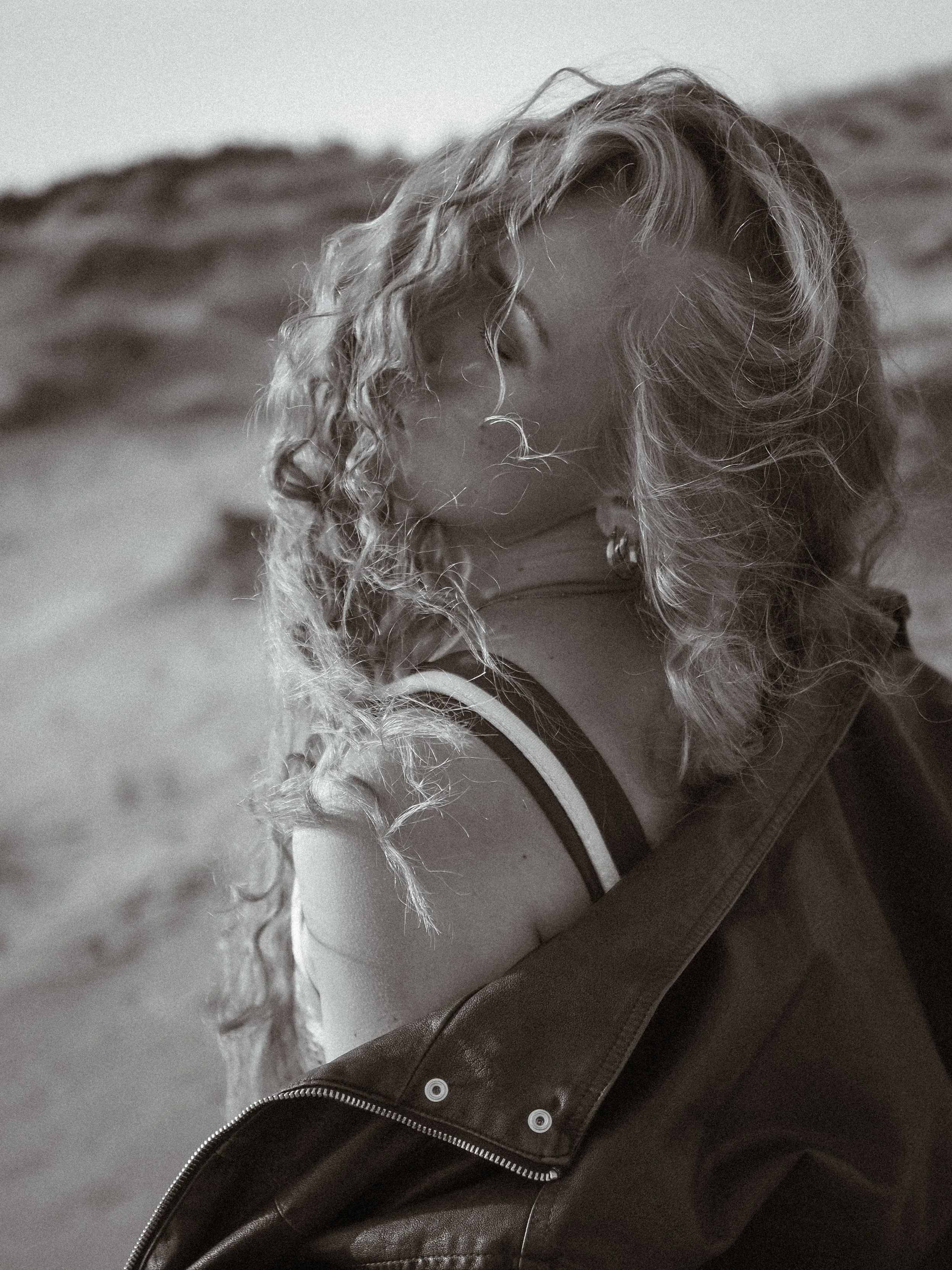 Vapaa-ajan nuori nainen, jolla on kihara, vaaleanruskea hiukset ja pukeutunut nahkatakki, istuu hiekkaisella rannalla.