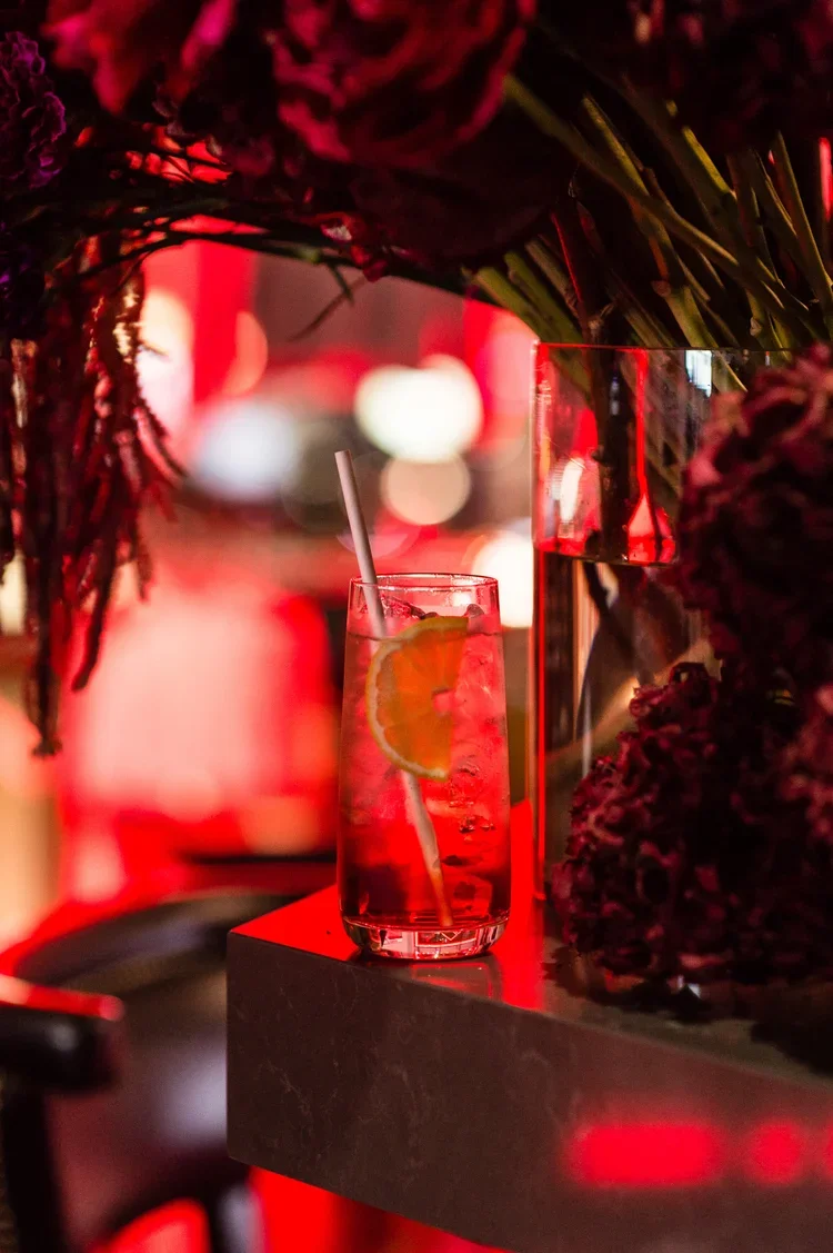 A glass of iced beverage with a slice of lemon and a straw on a black surface, surrounded by dark flowers, with blurred red and white lights in the background.