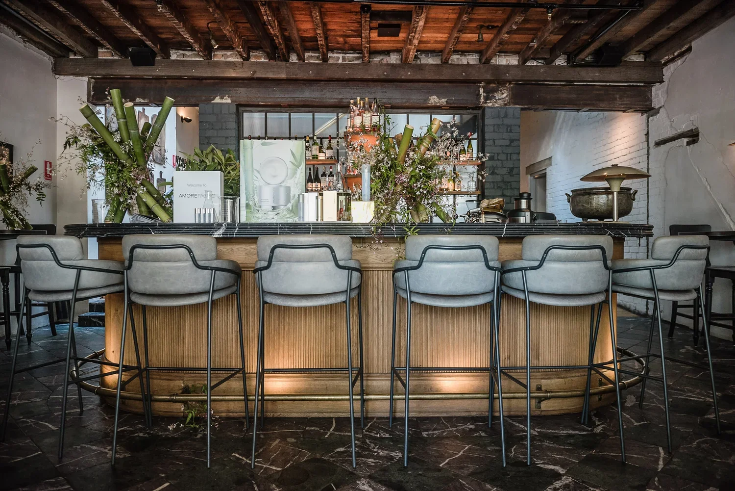 A modern bar with a marble top and beige upholstered bar stools with black metal frames. There are floral arrangements and bottles of alcohol behind the bar, with a decorative display of green bamboo stalks and greenery on either side. The ceiling ha