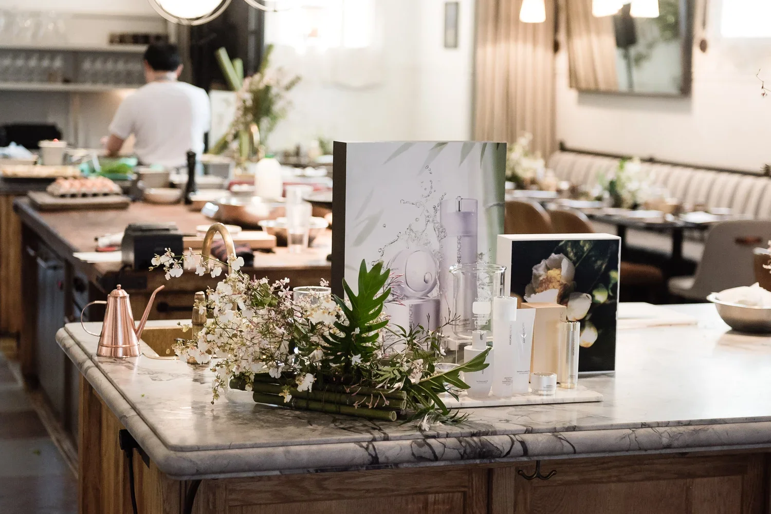 A marble-topped kitchen island decorated with flowers, skincare products, and promotional materials in a bright, modern dining space with a person in the background preparing food.