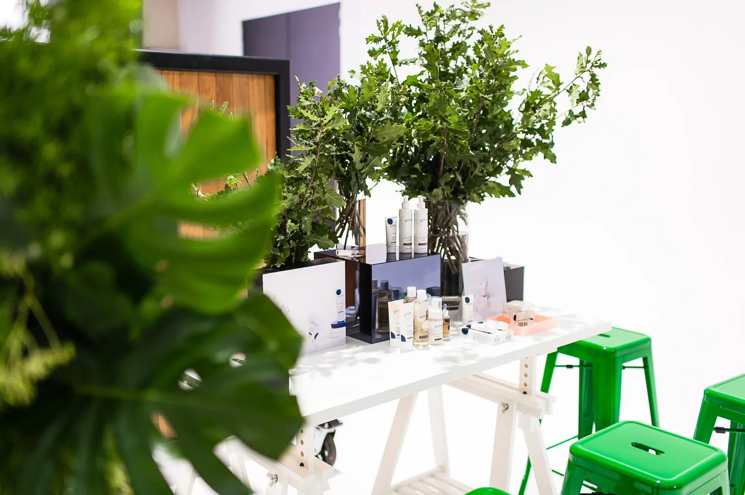A white table holding skincare or beauty products like bottles and tubes, with large green plants behind it, flanked by bright green stools, against a white background.