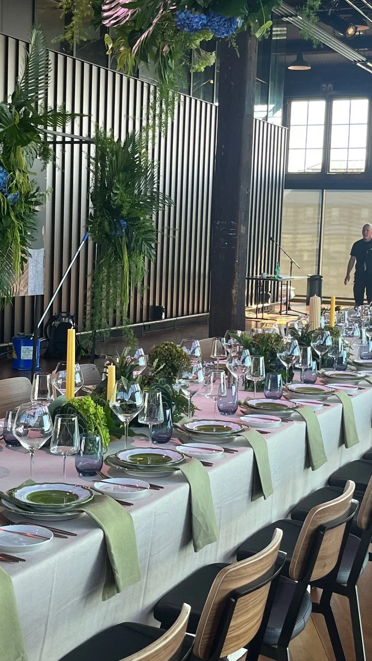 Long banquet table set with green napkins, glassware, and candles, decorated with green foliage and flowers in a modern, industrial-style venue with large windows and a stage in the background.