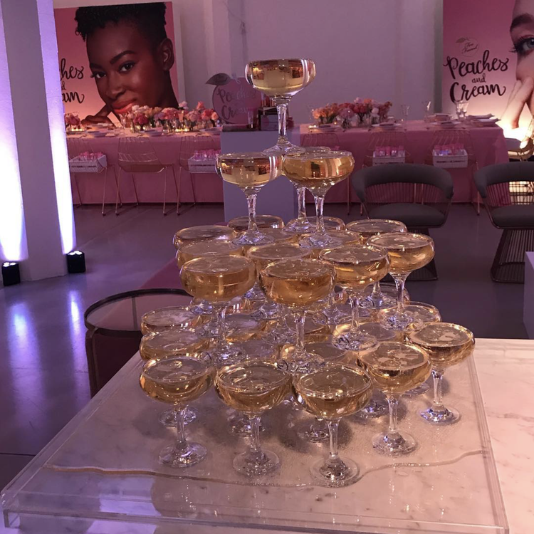 A pyramid of champagne glasses filled with a light golden beverage at an event decorated with pink-themed banners, flowers, and lounge seating.
