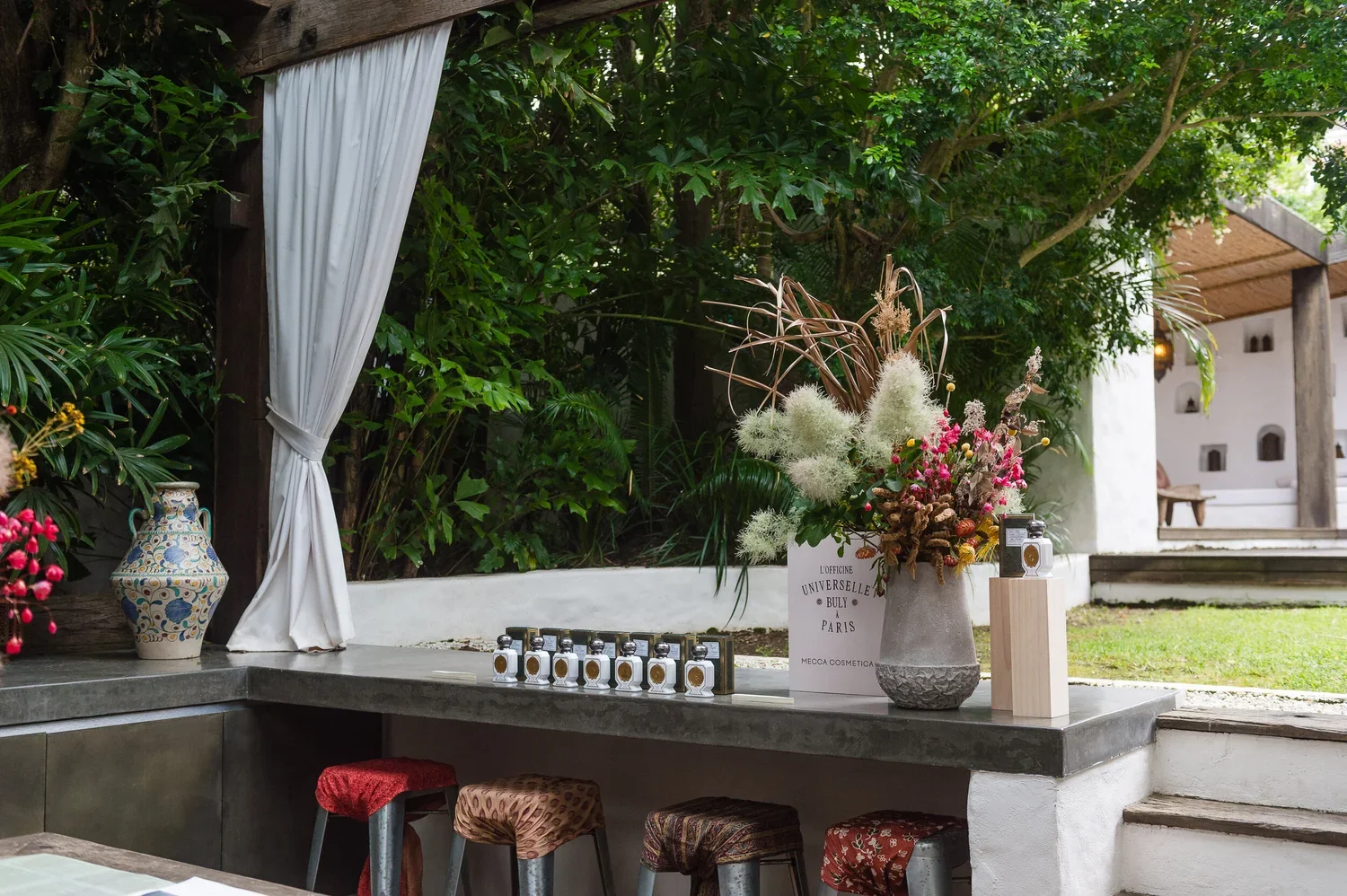 A decorated outdoor space with a large floral arrangement, small perfume bottles, a colorful vase, and stools with patterned cushions, set against lush greenery and a white building with steps and a porch.
