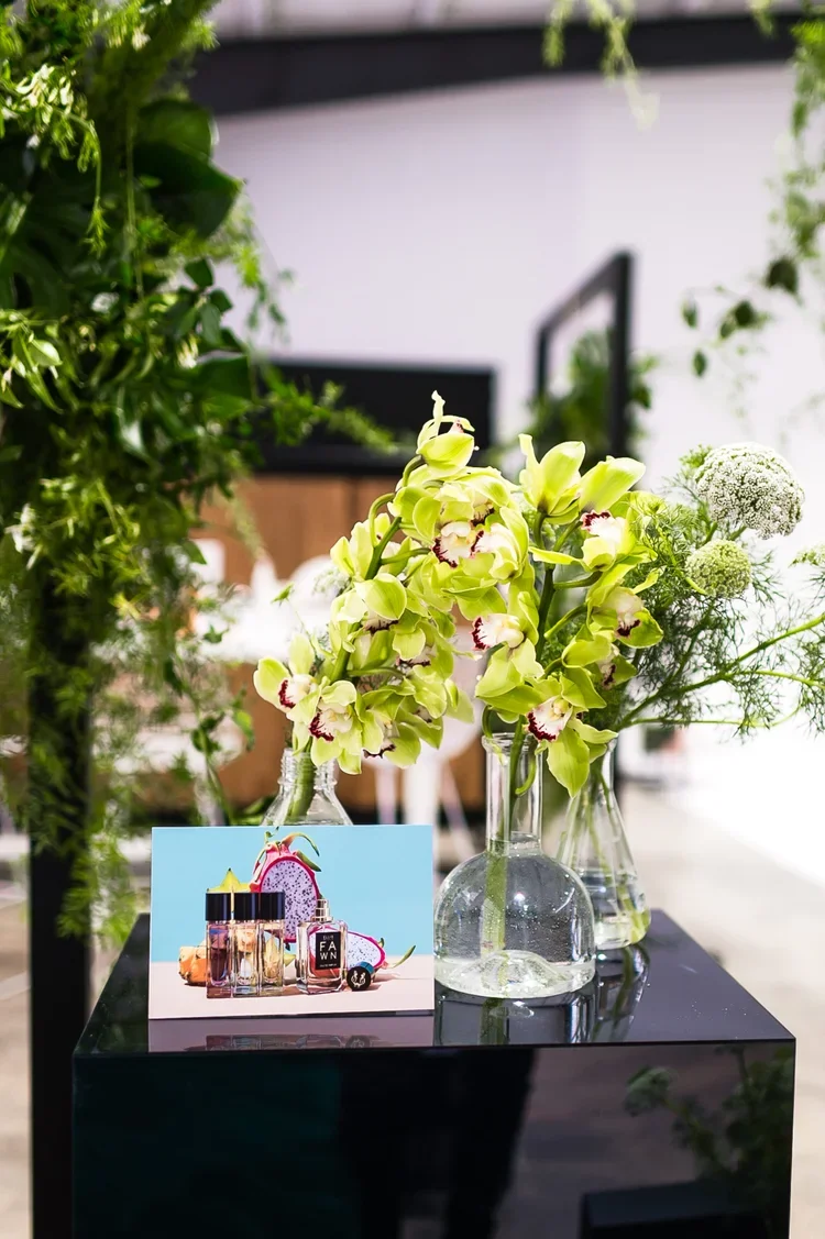 Vase with light green orchids and white flowers on a black table, with a perfume sample card and some bottles nearby.