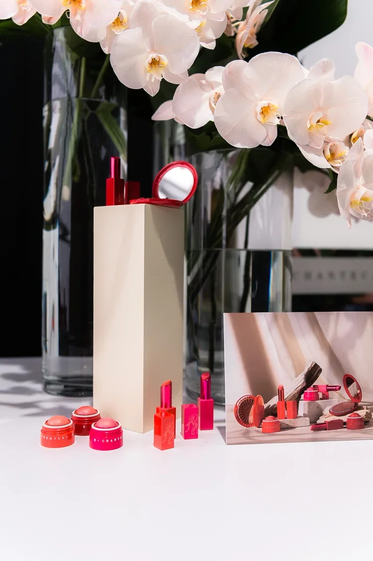 Display of various pink and red cosmetics, including lipsticks and round containers, placed on a white surface with tall glass vases of white orchids in the background.