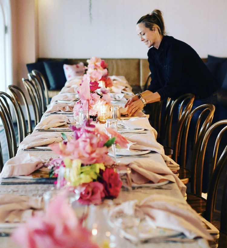 A woman setting a dinner table decorated with pink flowers, napkins, and glassware in a well-lit room.
