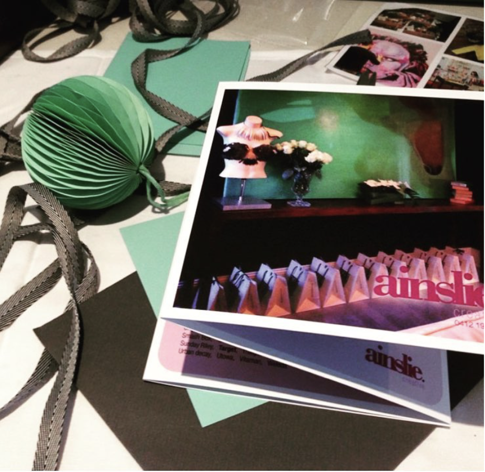 Photo of event invitations or tickets with a dark background, featuring images of stage lighting and audience, the word 'aainsic' in pink, and a visible subheading. The table also has scattered photographs, a black paper, teal cards, gray ribbon, and