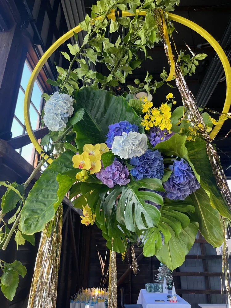 Decorative floral arrangement with blue, purple, white, and yellow flowers, large green leaves, and gold and yellow circular and strand decorations inside a building with large windows.