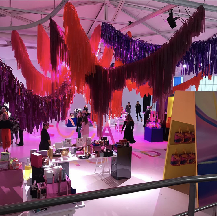 A colorful indoor event space decorated with hanging streamers in shades of pink, purple, red, and orange. Several people are browsing booths with various products on display, illuminated by pink and purple lighting.