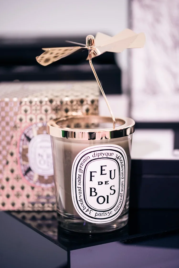A metallic candle holder with a white label and black text, shaped like a perfume bottle, topped with a gold and white paper pinwheel. In the background, there are patterned boxes and a blurred interior setting.