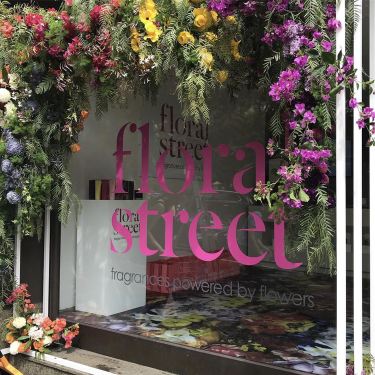 A store window decorated with colorful flowers including yellow, purple, and white blooms, with the words 'flora street' displayed prominently on the glass in pink and black lettering. Inside the store, floral arrangements and a white counter are vis