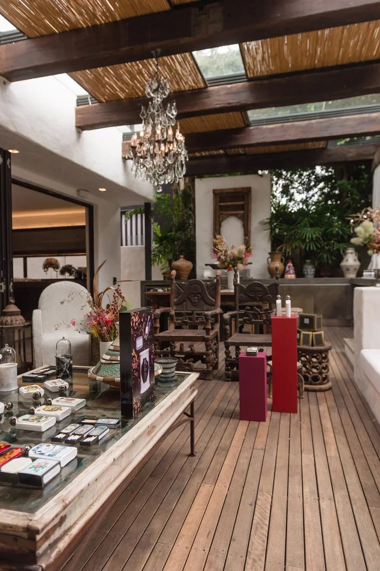 An interior space with wooden flooring, a chandelier hanging from a thatched ceiling, and various decorative items including vases, flowers, and ornate chairs.